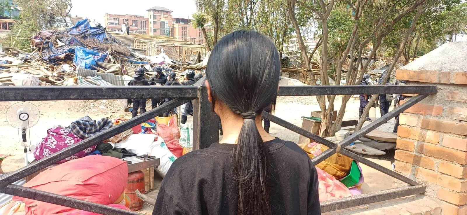 Ein Mädchen mit langem Zopf steht mit dem Rücken zur Kamera vor den Trümmern des geräumten Slums in Thapathali, Kathmandu, und blickt auf die Reste der Hütten.