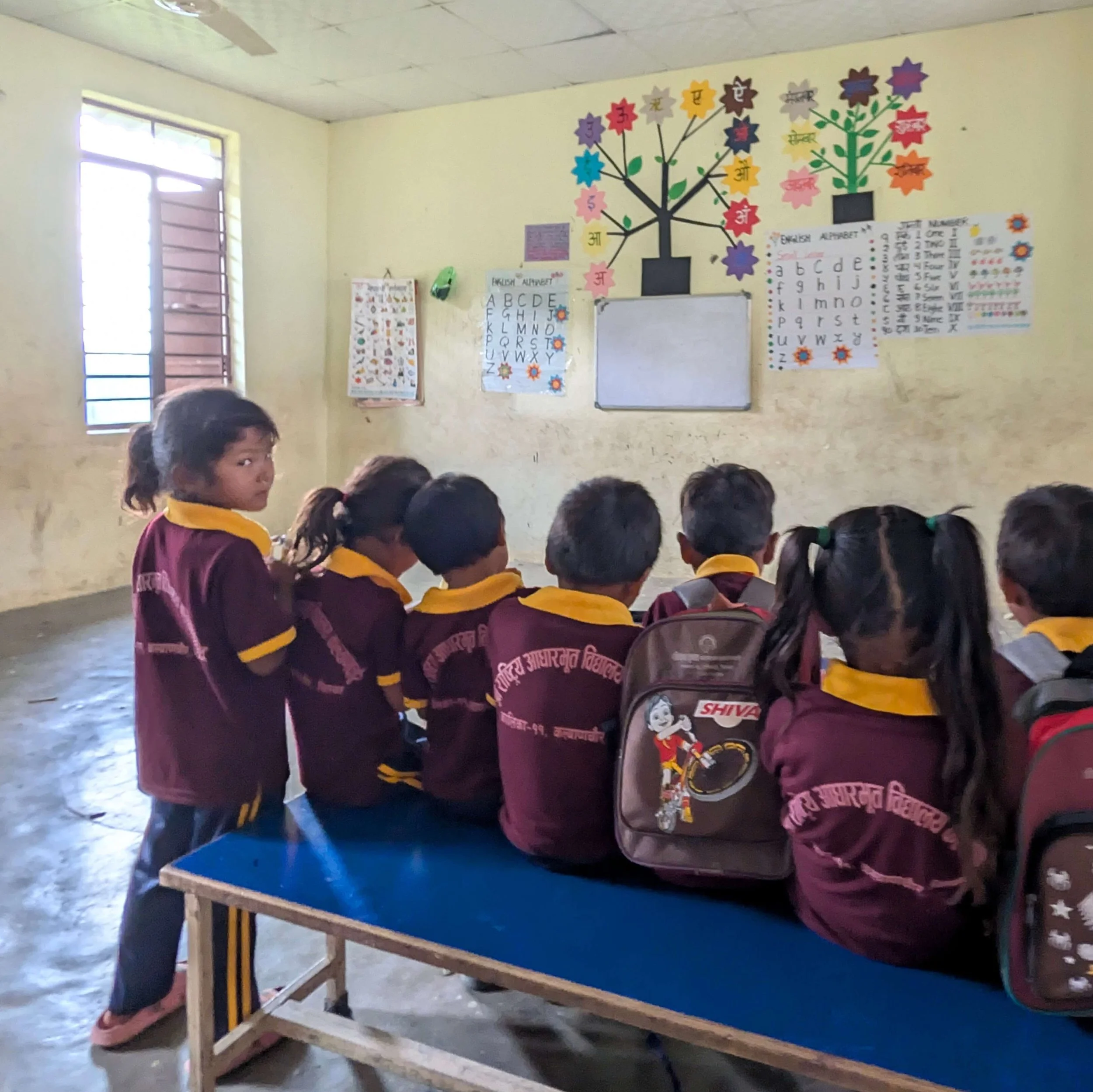 Schulkinder in weinroten Uniformen sitzen mit Schulranzen von hinten auf einer Bank im Klassenzimmer der Bergschule in Chepang, Chitwan, Nepal, an der Wand Alphabet-Poster auf Nepali und Englisch, ein Mädchen blickt zurück