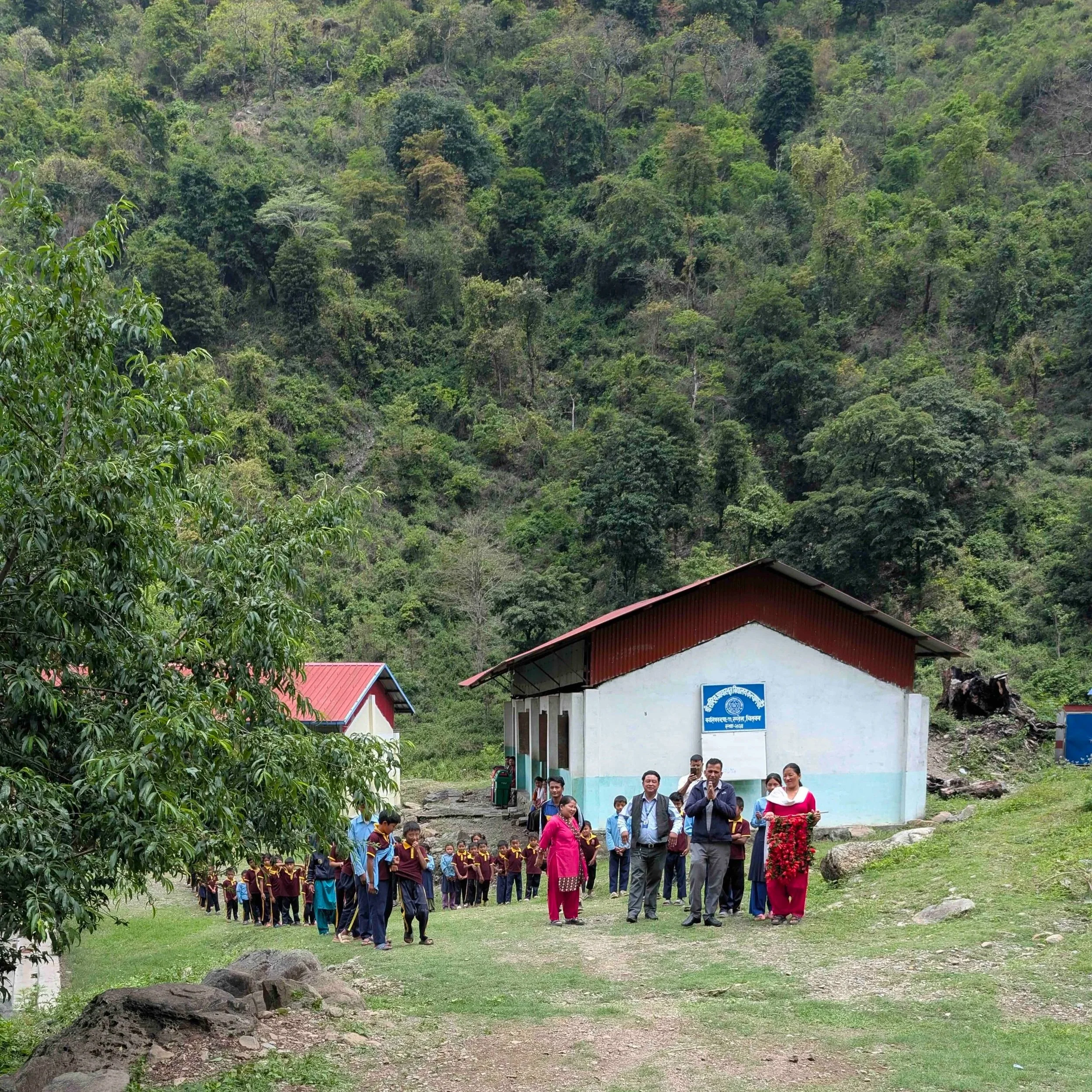 Chepang-Schulkinder stehen in Reihen vor ihrer Schule in den Hügeln von Chitwan, Nepal, dahinter ein steiler bewaldeter Hang