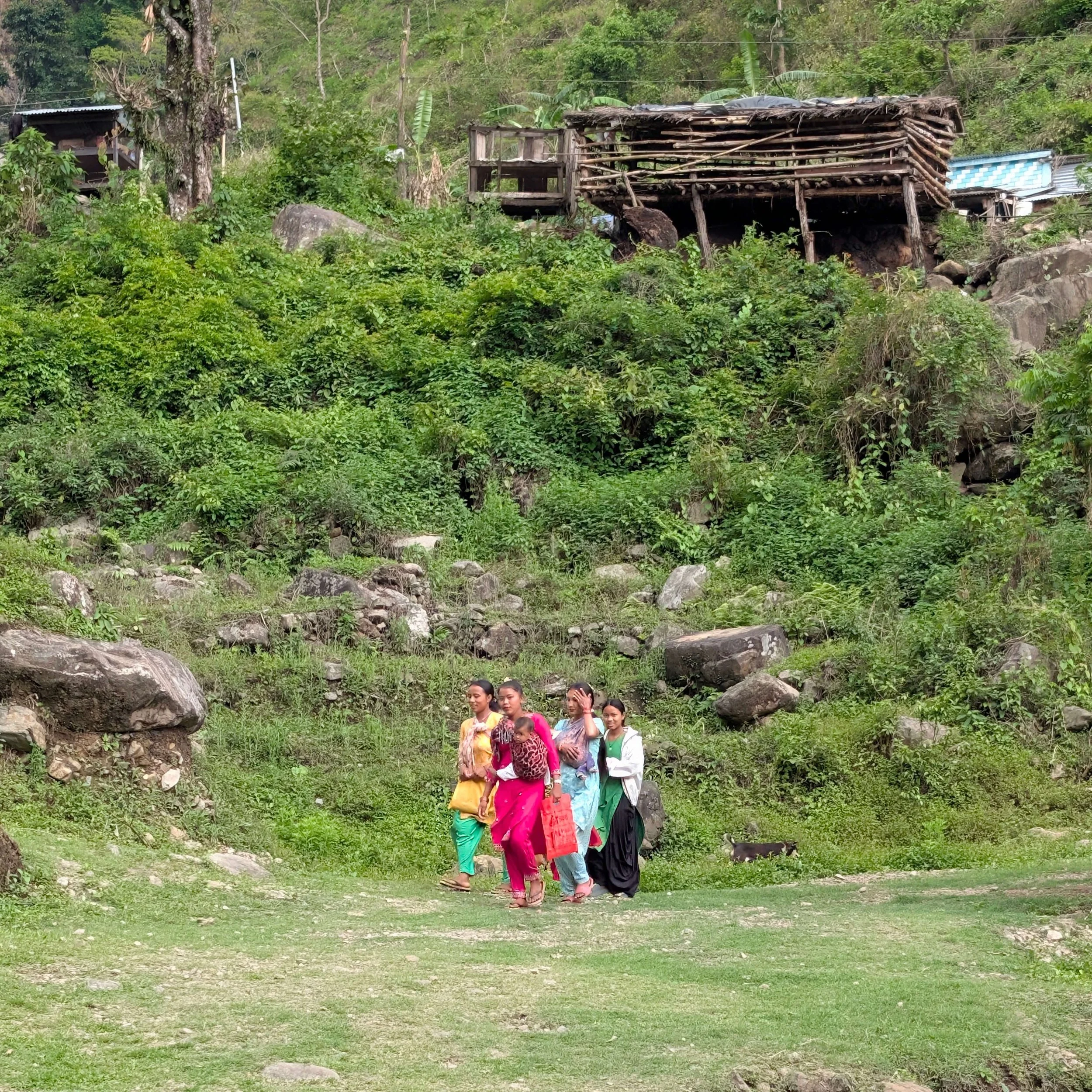 Junge Frauen in bunten Kleidern gehen mit Babys in Tragetüchern einen Bergpfad entlang. Im Hintergrund sind marode Holzhäuser zu sehen.