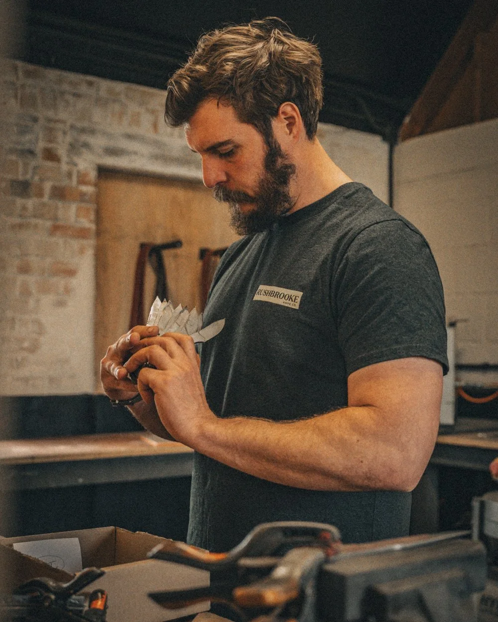 Man with a beard and dark hair looking down and holding a small paper with the letters 'RUSHBROOKE' on his chest, working in a workshop with brick wall and tools in the background.