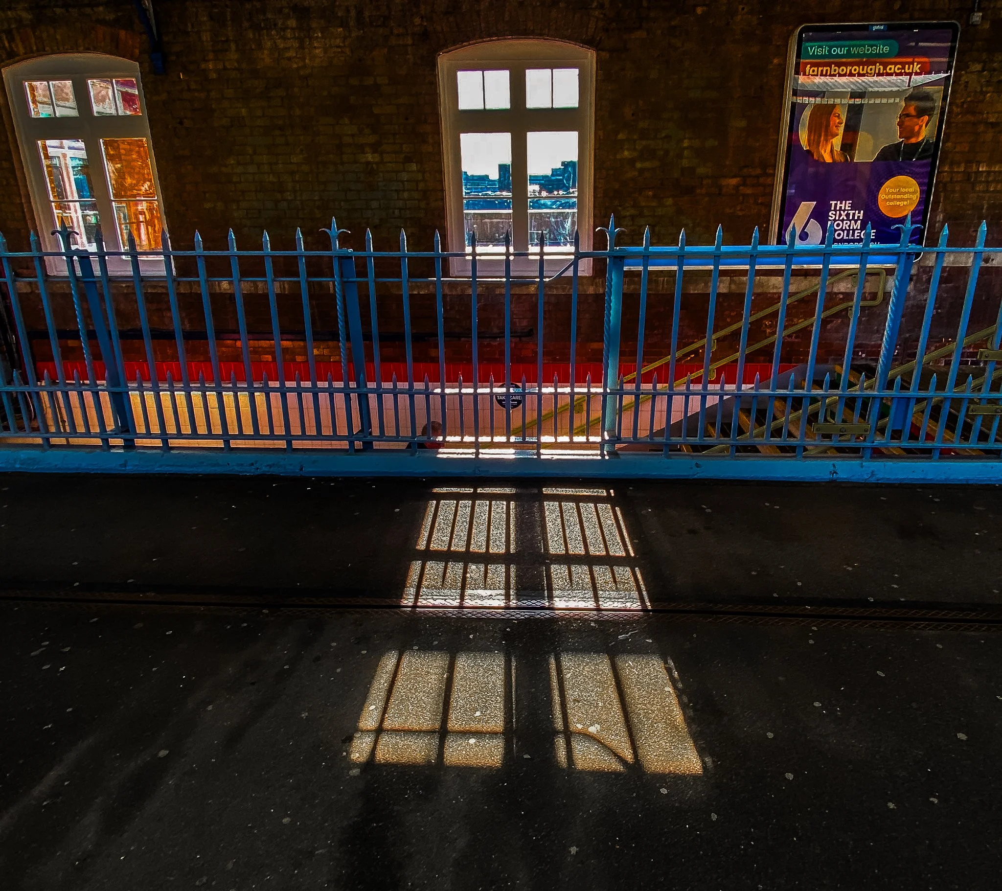 Sunlight casting a shadow of a blue fence and windows on the ground inside a brick building with two large windows, a staircase, and a large digital advertisement display.