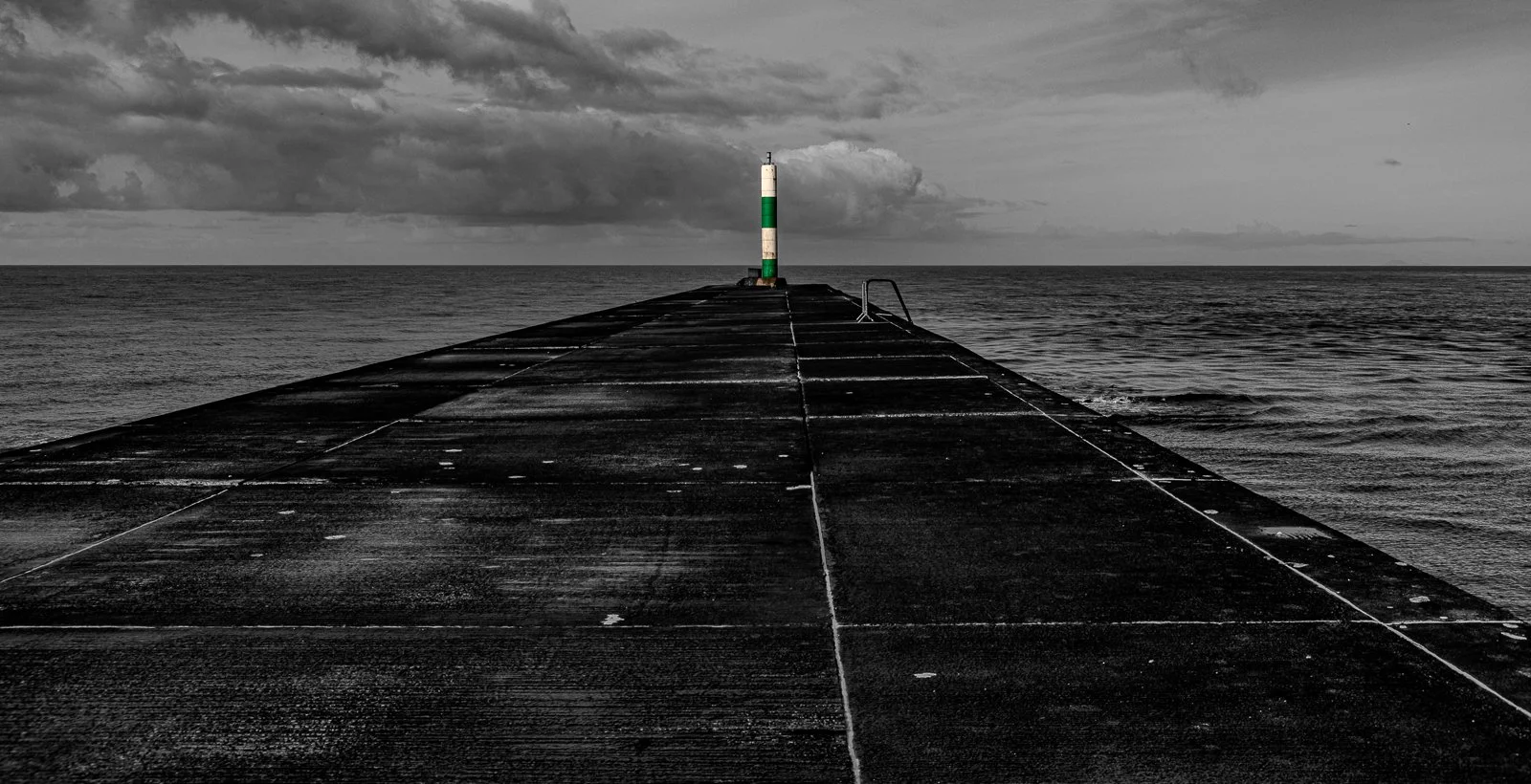 A black-and-white photo of a concrete pier extending into the ocean, with a striped lighthouse at the end of the pier and stormy clouds in the sky.