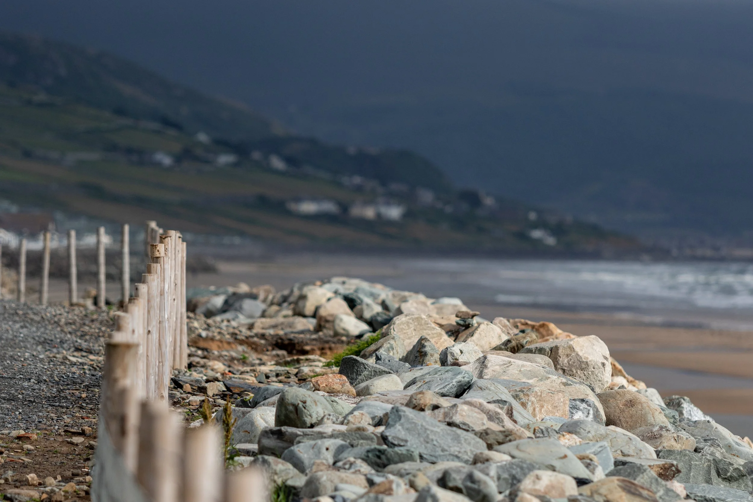 Rocky shoreline with a weathered wooden fence in the foreground, and hills with scattered houses in the background, under a cloudy sky.