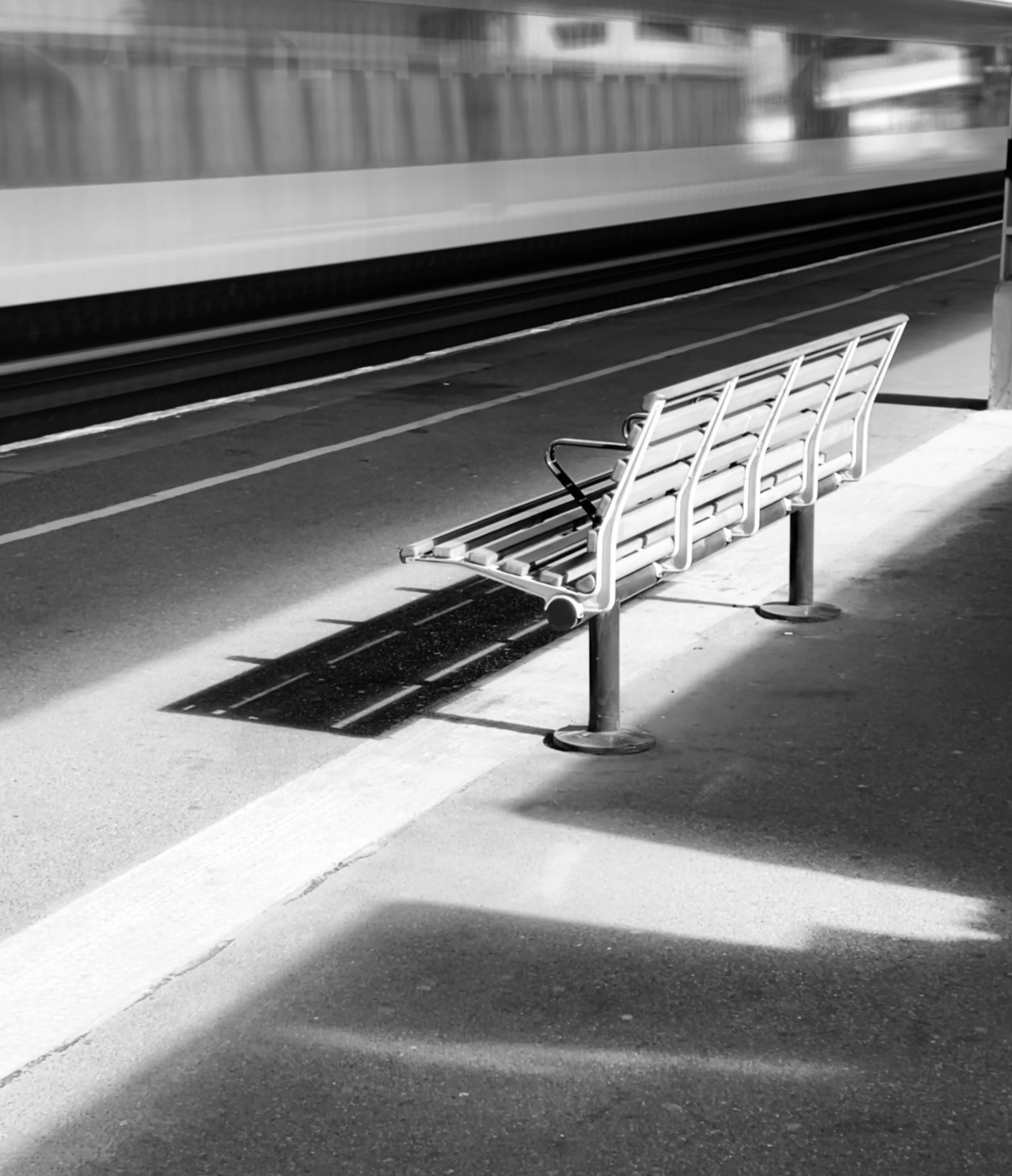 Empty train station platform with a metal bench and a moving train blurred in the background, casting a shadow on the pavement in black and white.