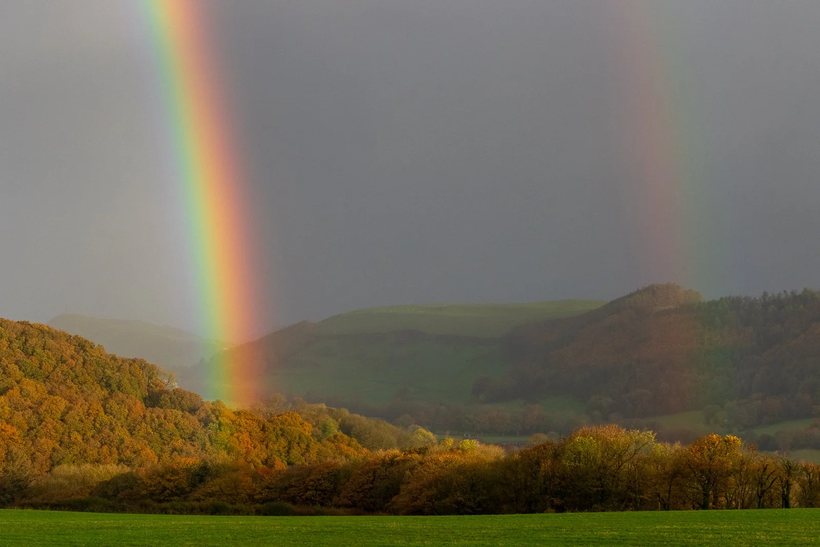 A double rainbow over a landscape with green hills and trees in fall colors.