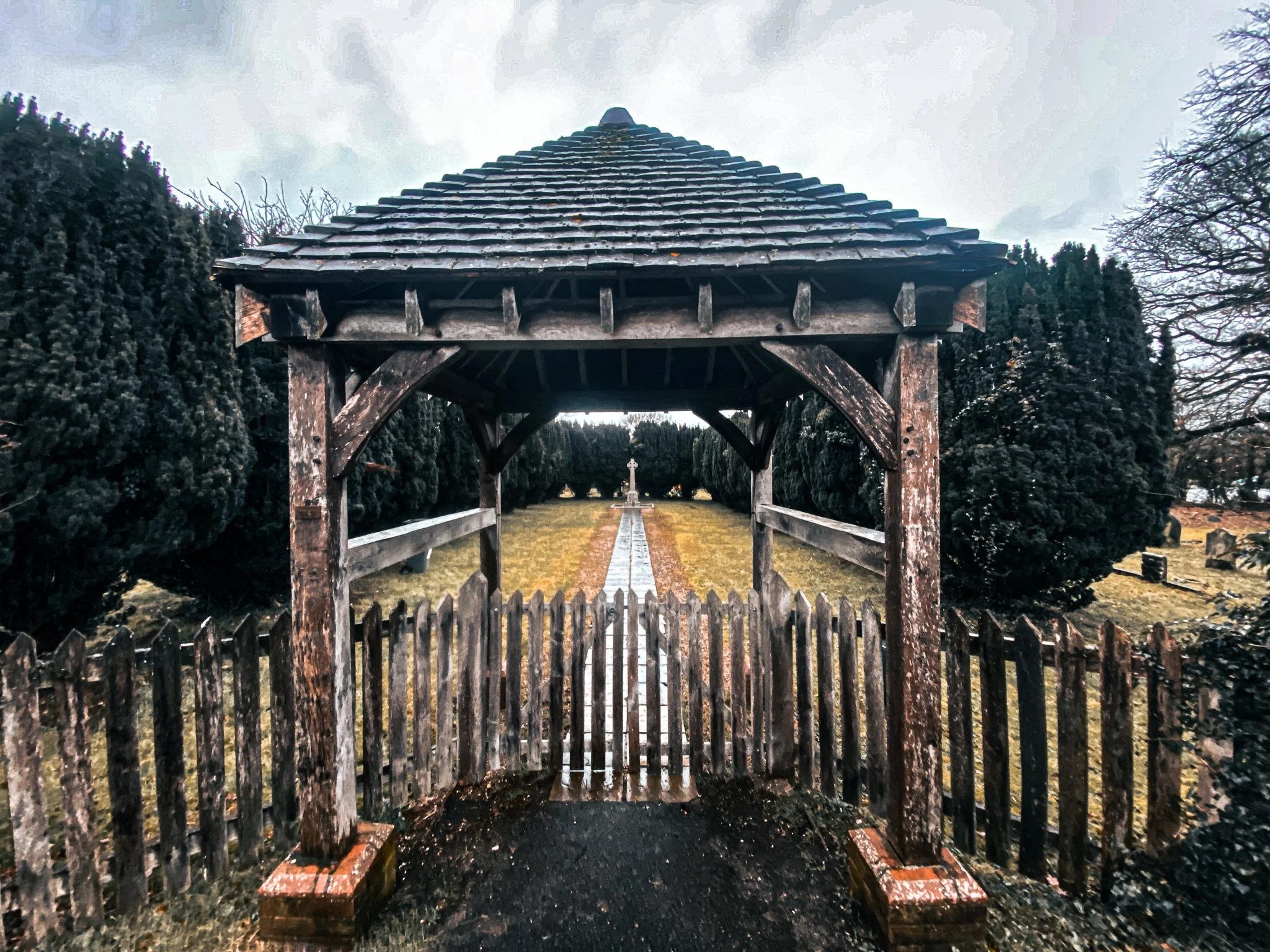A wooden gazebo with a shingled roof at the entrance of a walking path leading to a cross monument in a garden or cemetery, surrounded by trees and overcast sky.