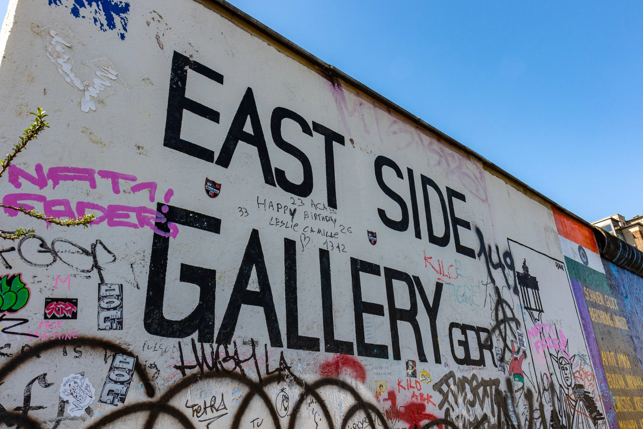 Graffiti-covered wall with large text saying 'EAST SIDE GALLERY' and various smaller graffiti, stickers, and drawings.
