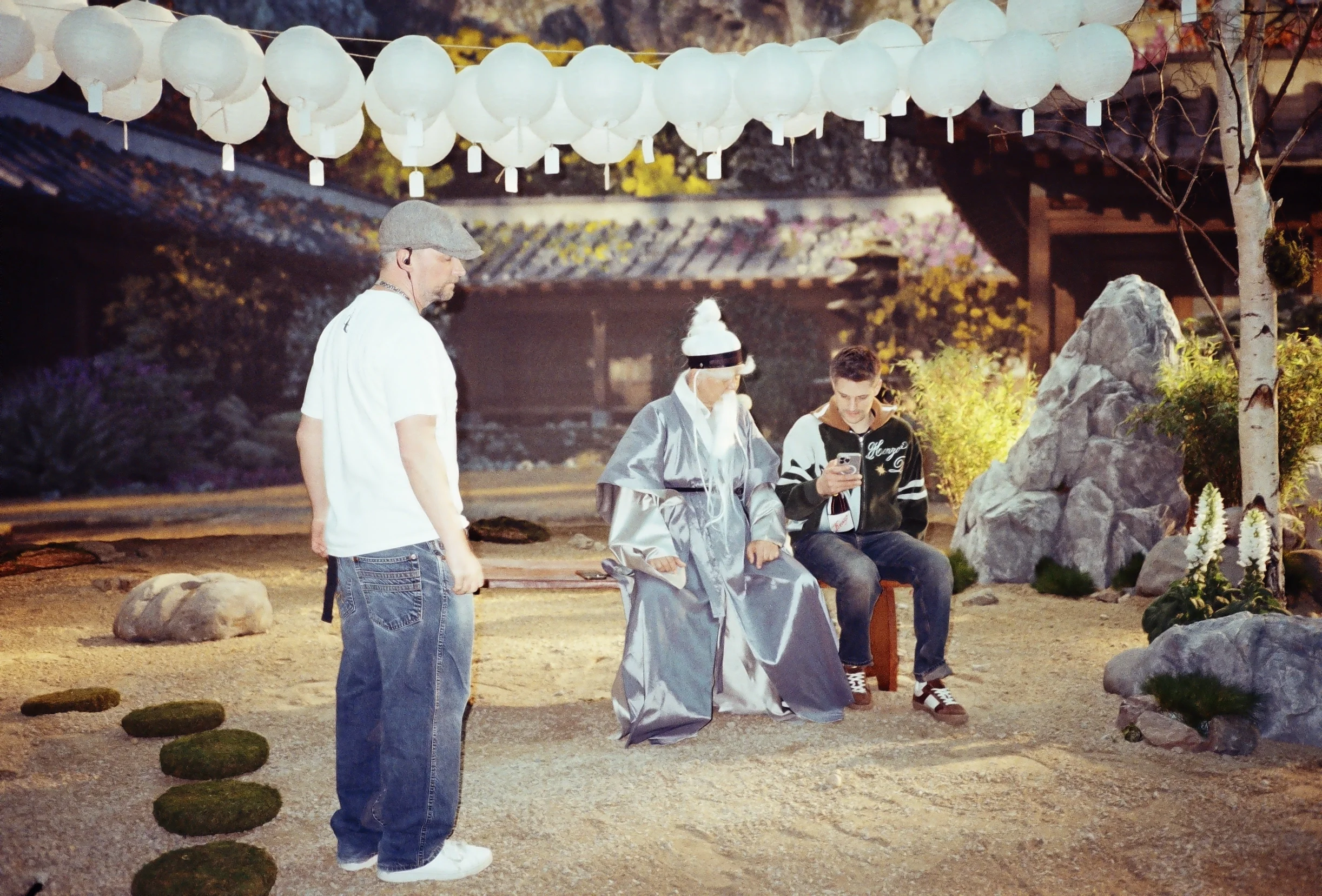 Japanese tradition ceremony with people in traditional attire, standing around a rock formation and lanterns hanging overhead, outdoors in evening.