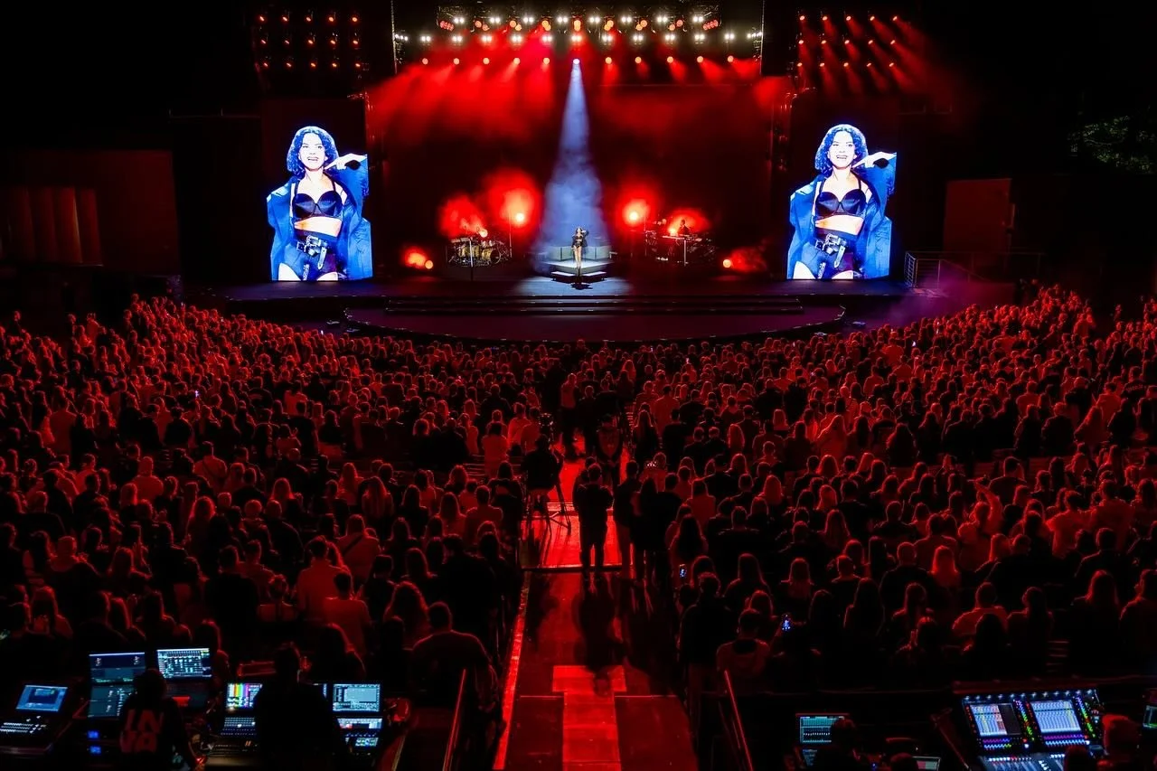 A large audience watching a concert on an outdoor stage with bright red lighting and large screens displaying a female performer in a black outfit.