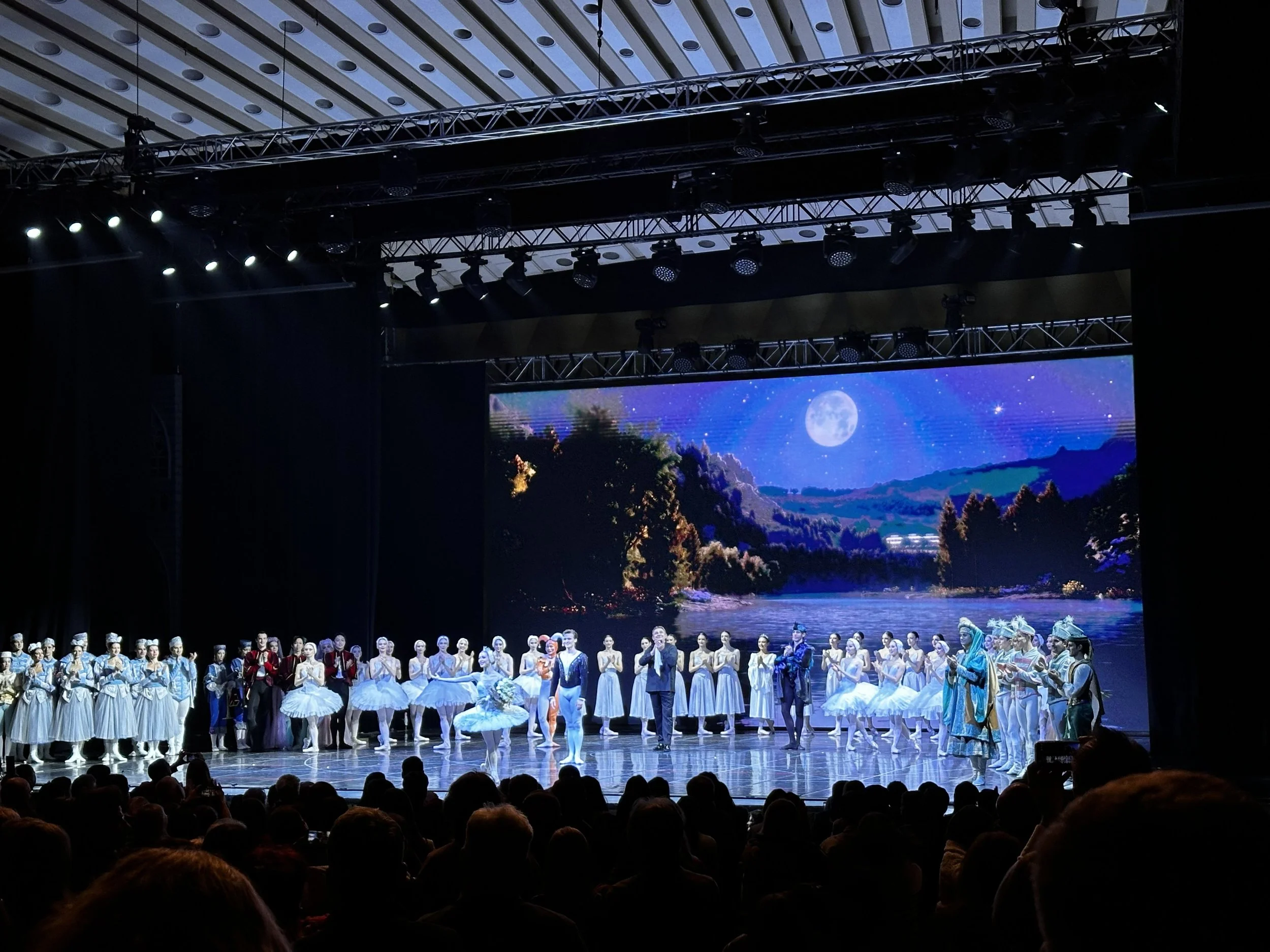 A stage with performers in historical costumes, a scenic backdrop of mountains and a river, and stage lighting overhead.