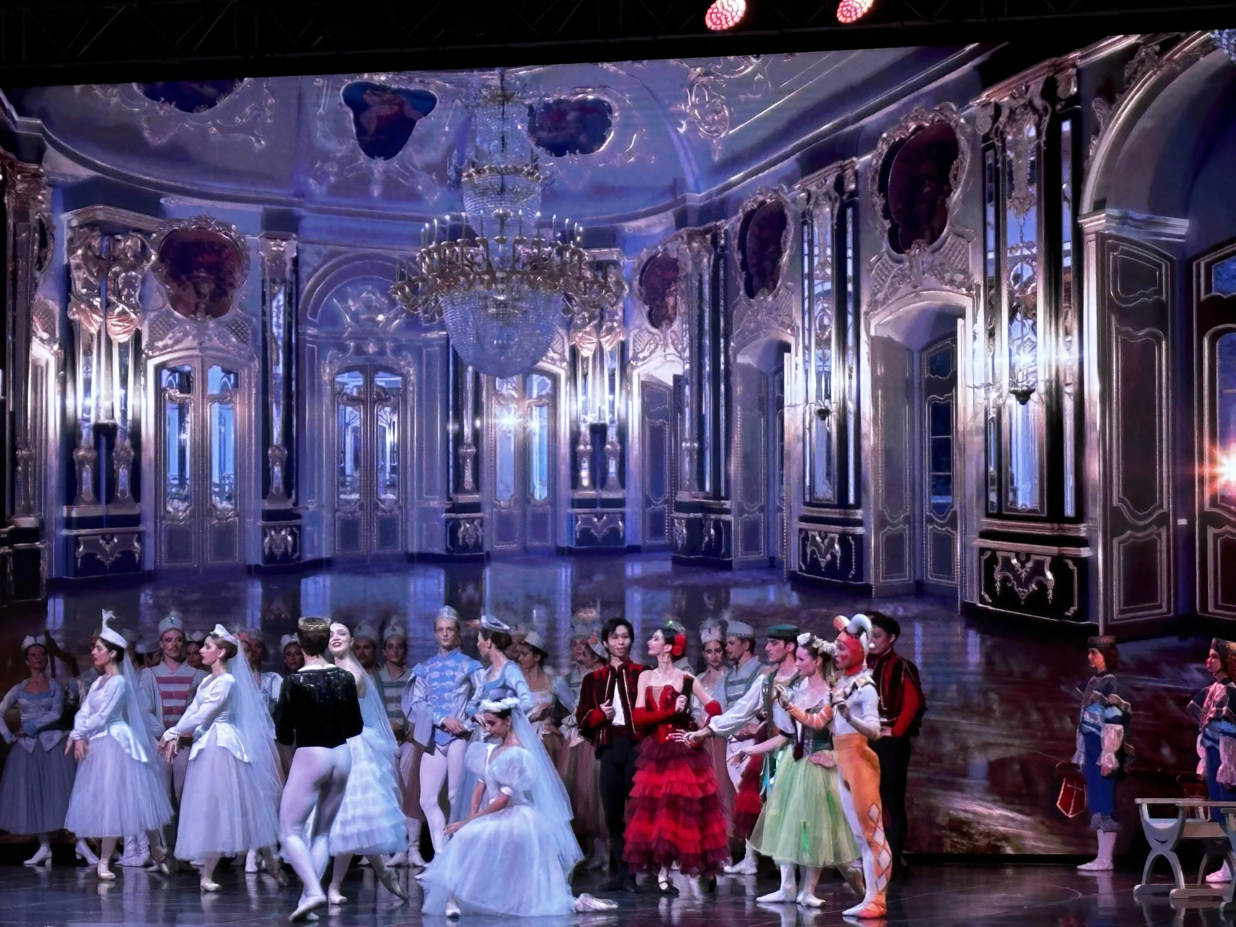 Ballet dancers dressed in costumes on stage, with a luxurious chandelier and ornate mirrors in the background, during a stage performance.