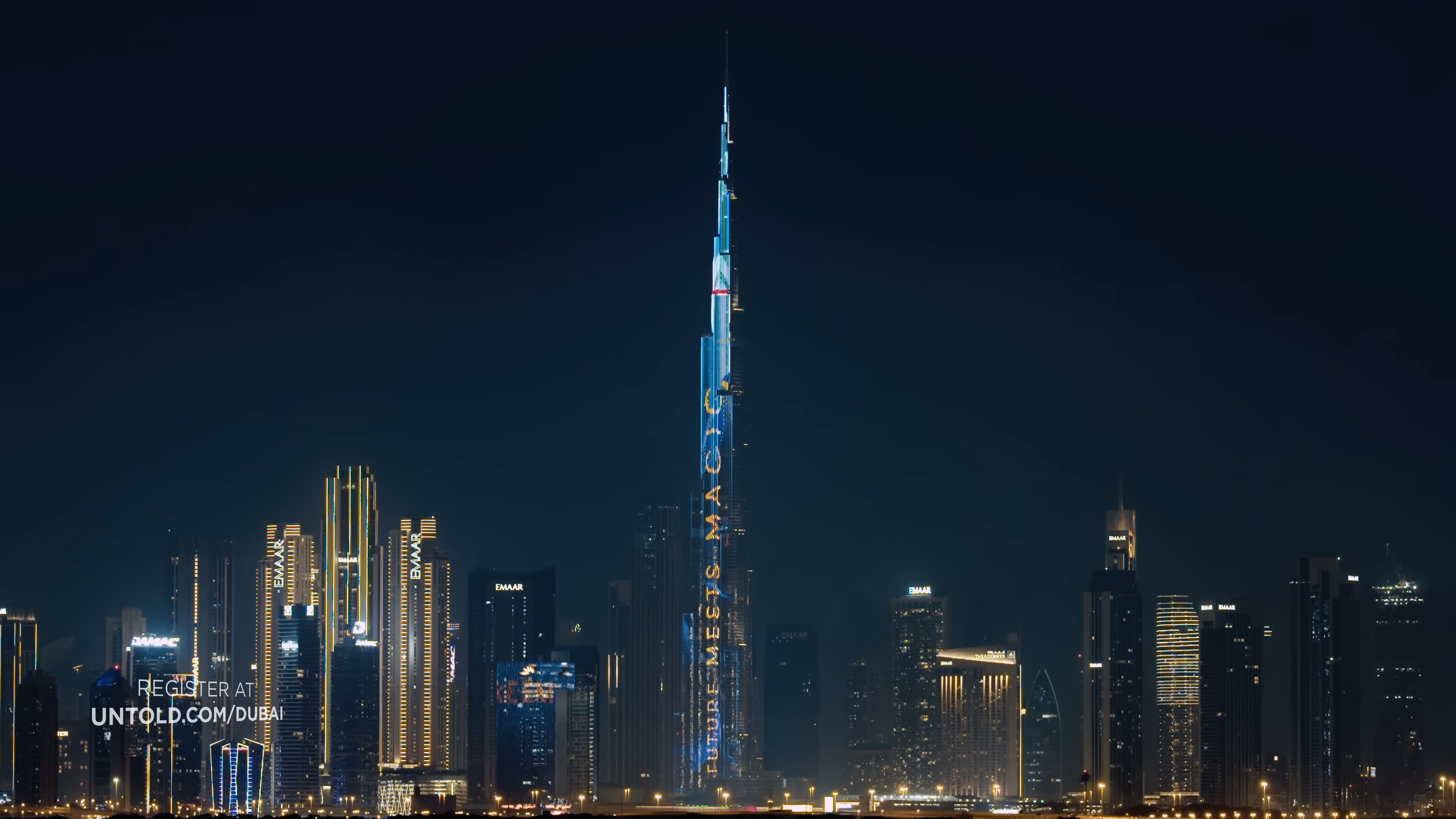 Nighttime city skyline featuring the tallest building in Dubai, illuminated with blue and white lights, with other high-rise buildings surrounding it.