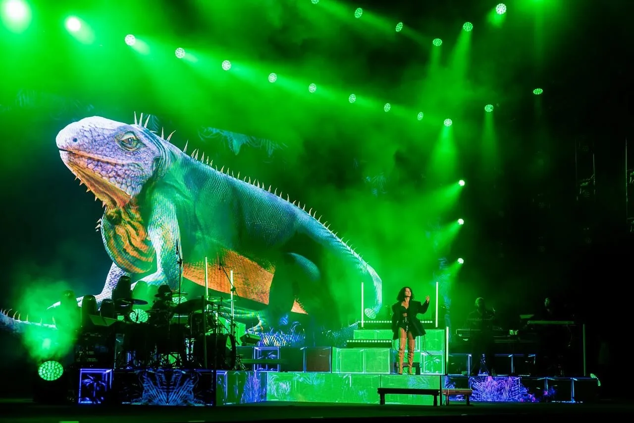 A stage with a large digital projection of a colorful iguana, surrounded by green stage lights, with a singer and musicians performing.