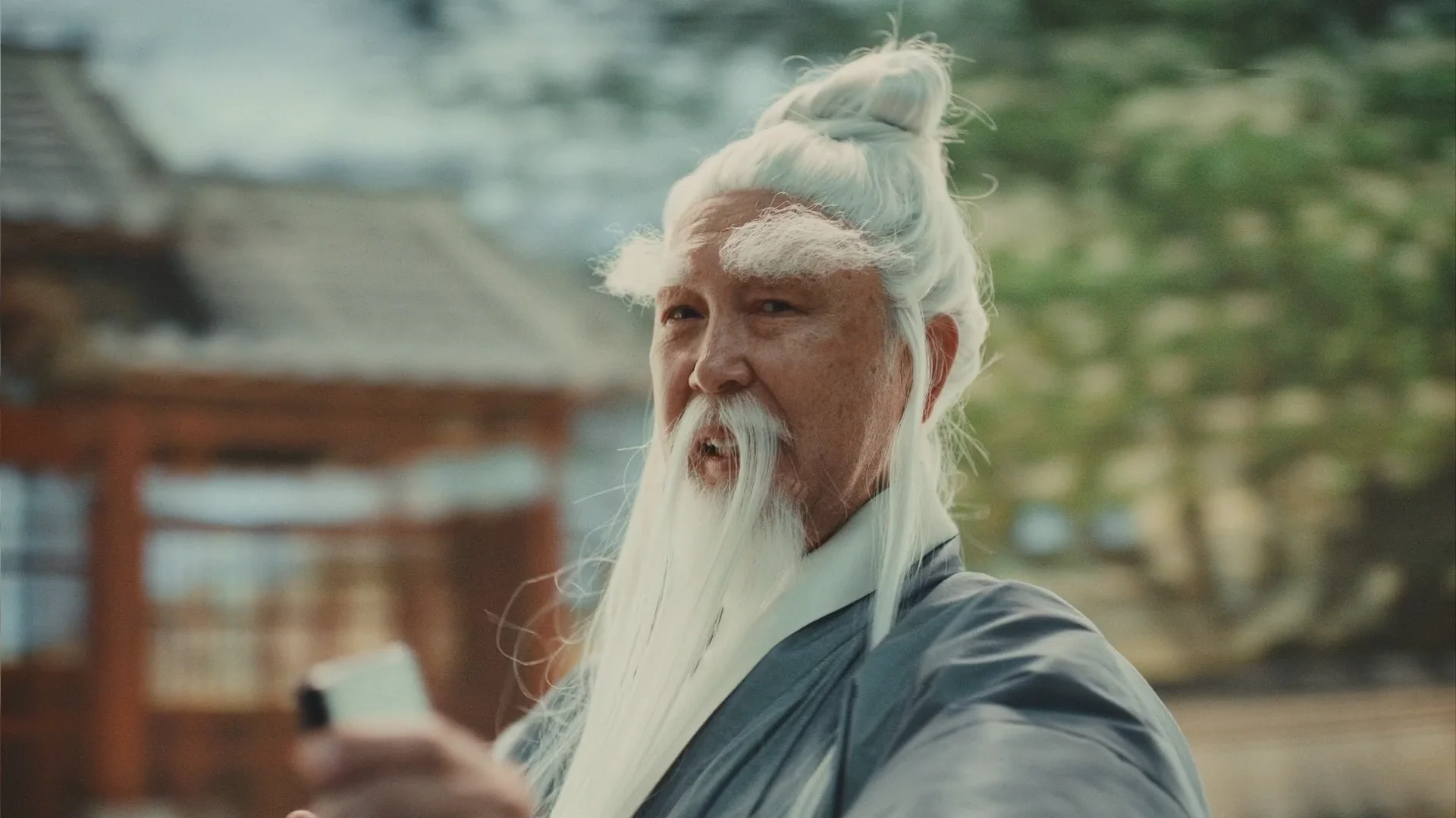 An elderly man with long white hair and a beard, dressed in traditional robes, pointing a gun with an intense expression.