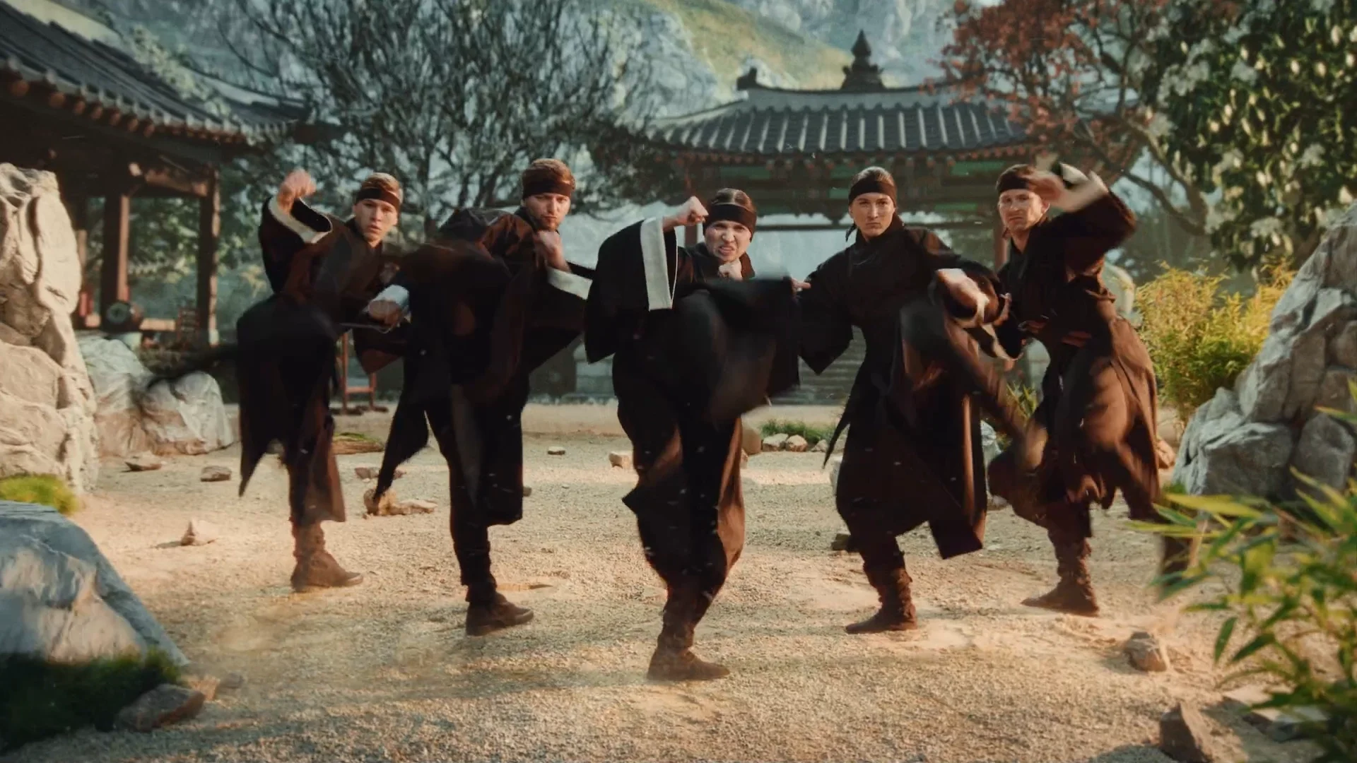 Group of people dressed in martial arts uniforms practicing martial arts outdoors in front of traditional Asian-style buildings with trees and rocks in the background.