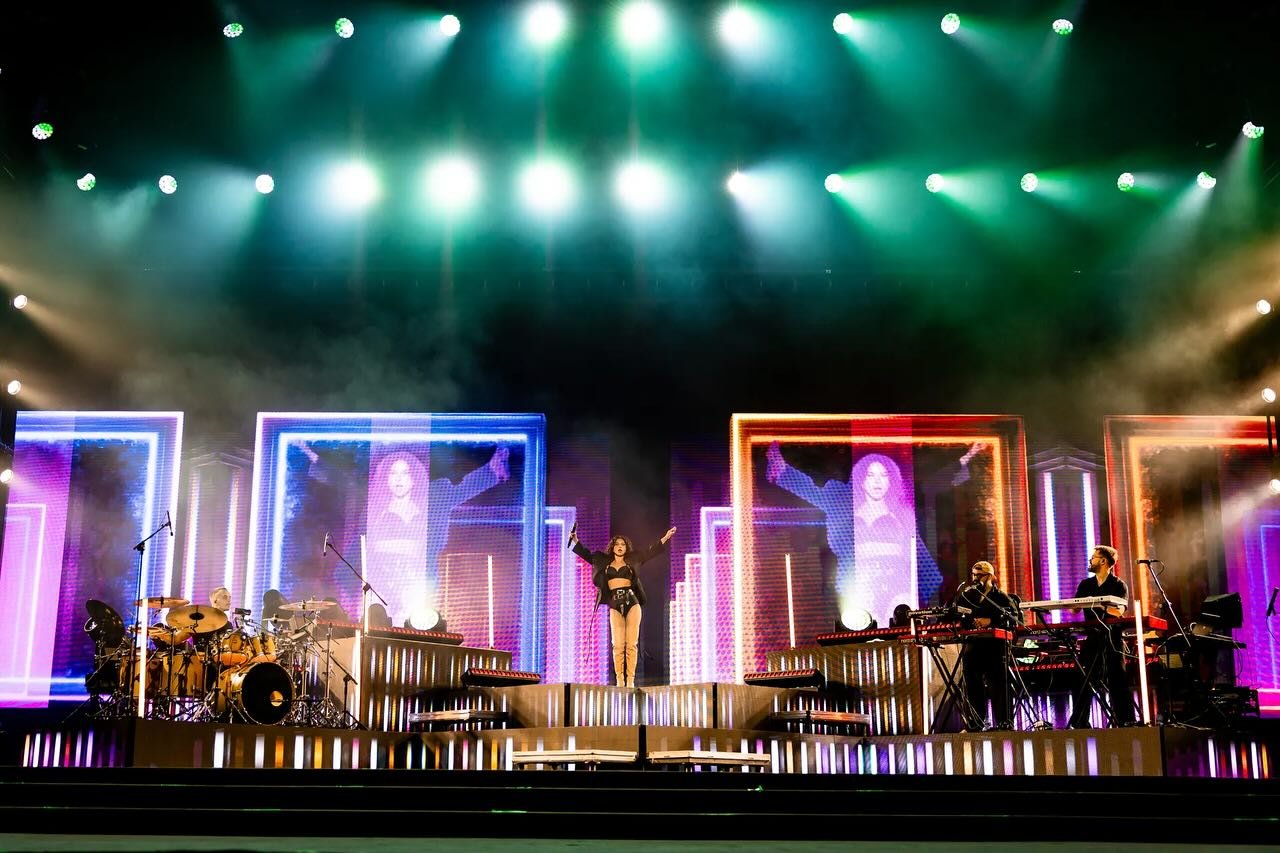 Performers on stage with colorful neon-lit backgrounds, including a singer at the center, drummers, and keyboardists, during a concert or music show.