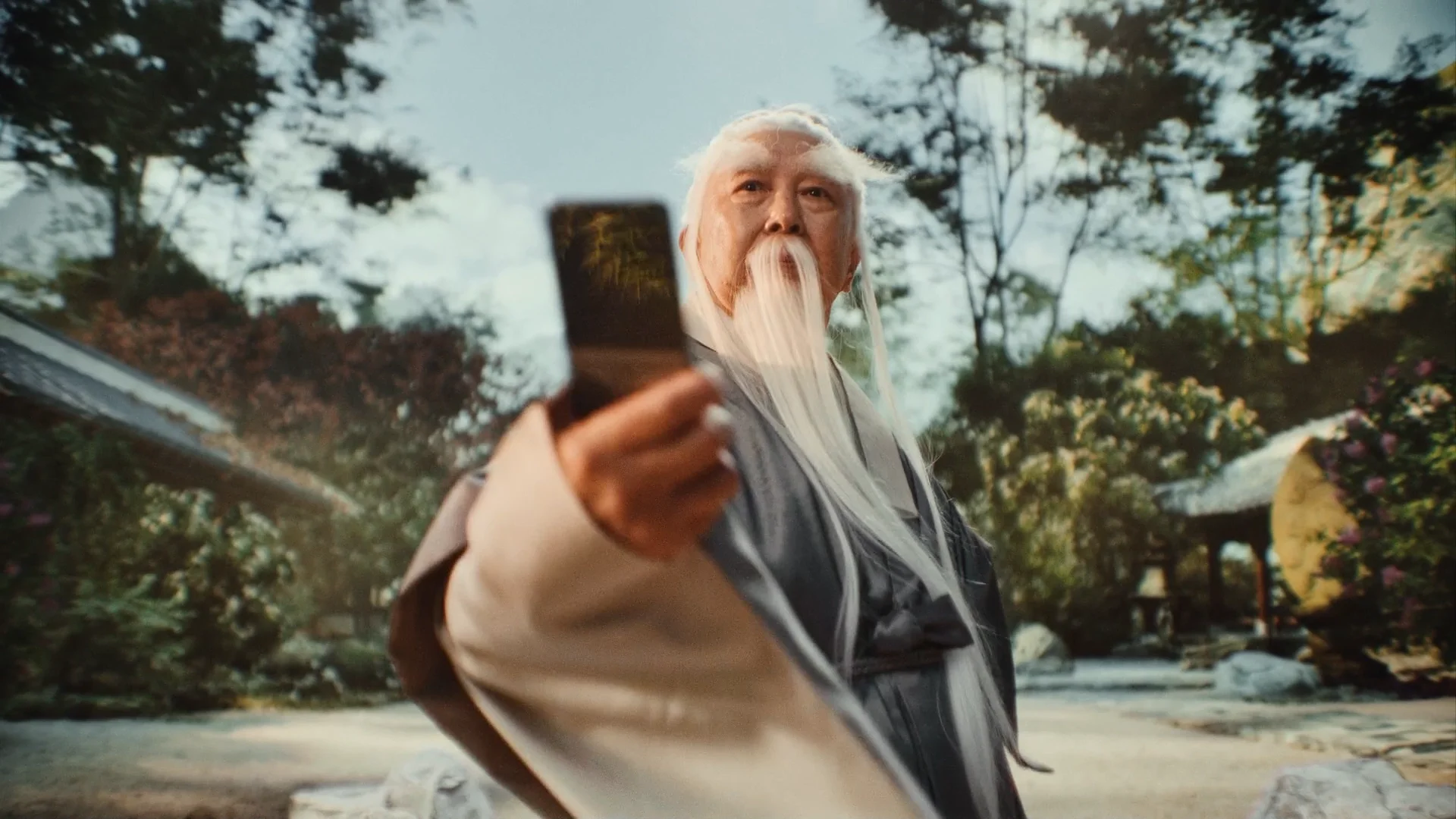 A person dressed as an elderly man with long white beard and hair, wearing traditional robes, is holding a smartphone and looking at the camera against an outdoor garden background with trees and a traditional structure.