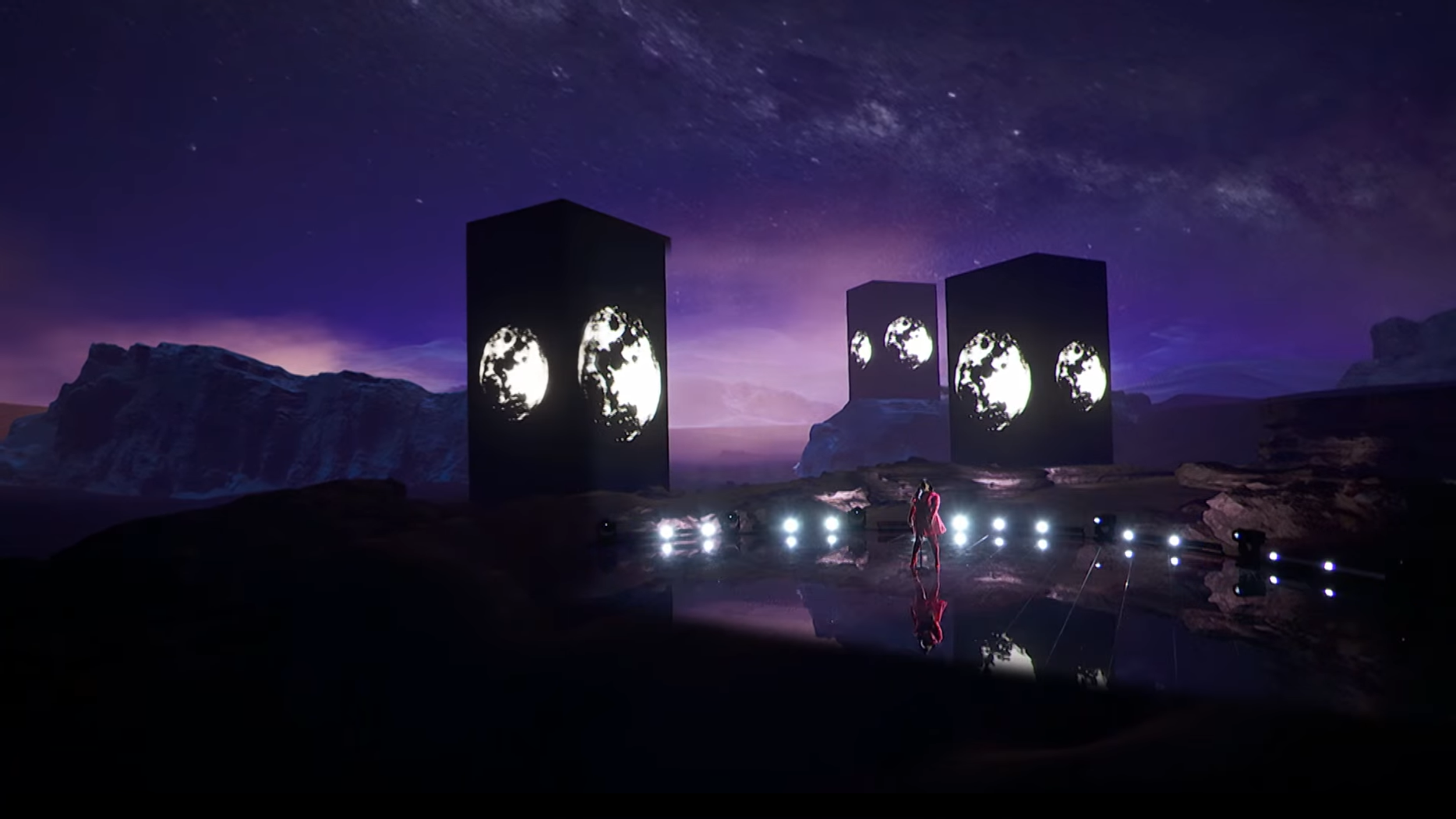 Nighttime outdoor scene with four tall rectangular screens displaying moon images, set against a mountainous landscape under a starry sky, with a person standing in front of bright lights reflecting on a wet surface.