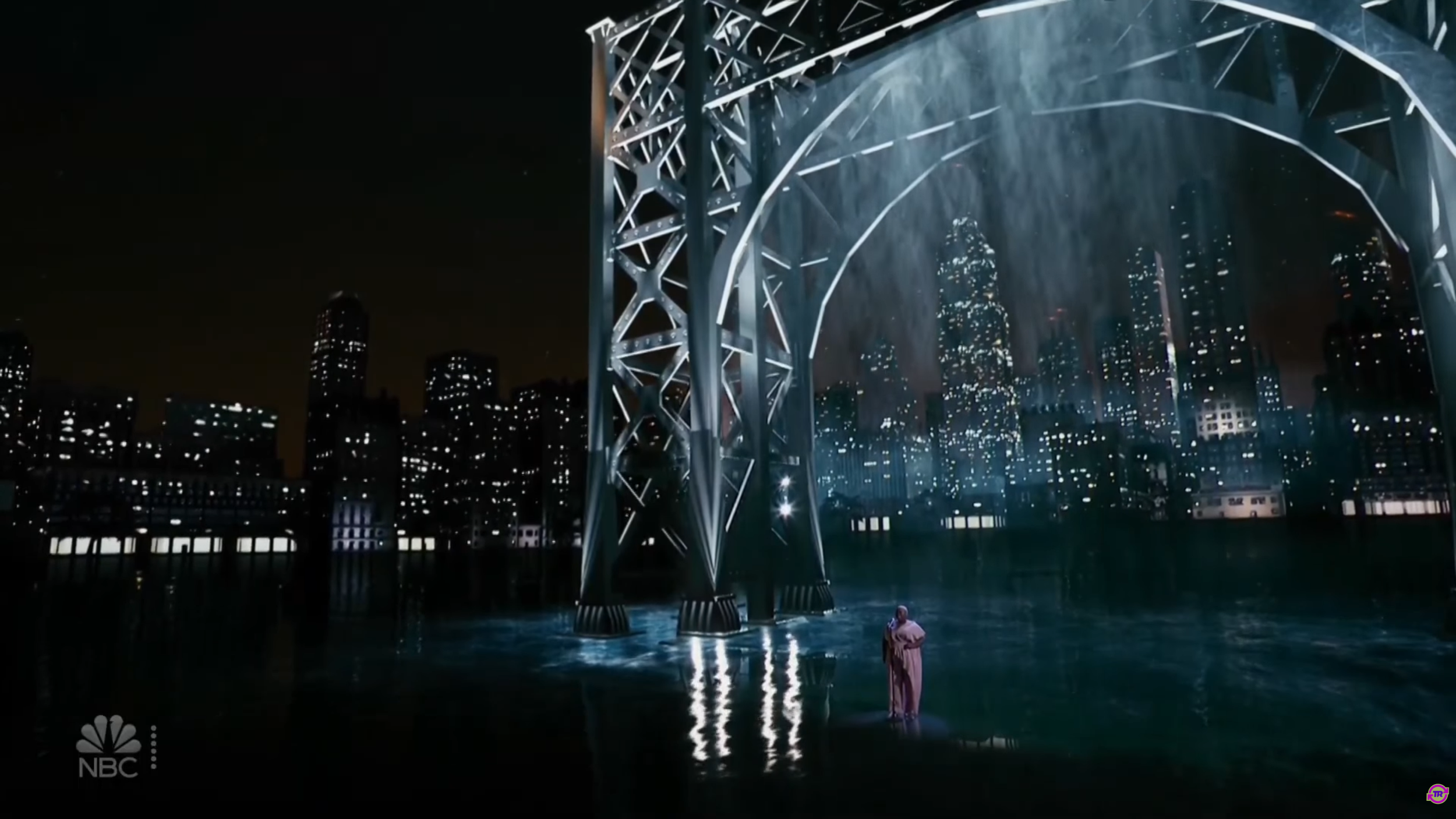 Night scene with a city skyline in the background, featuring tall illuminated buildings. In the foreground, there is a large, metallic, geometric structure with water flowing over it, creating a waterfall effect. A person stands on the water near the