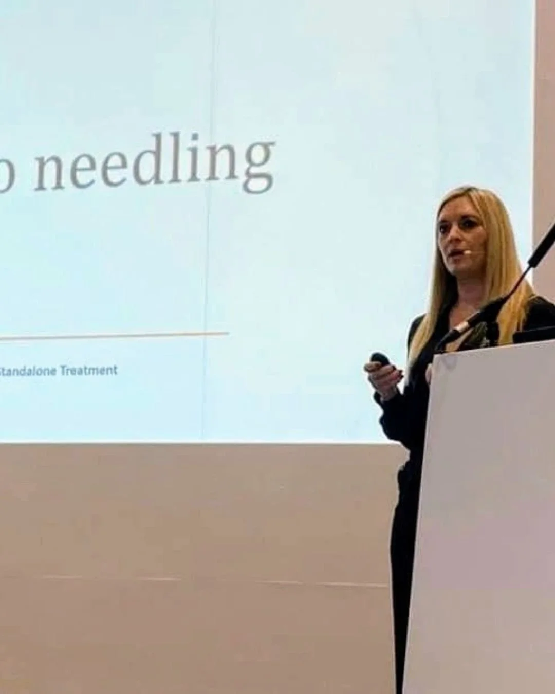 A woman with long blonde hair giving a presentation, standing next to a large projection screen with partially visible text.