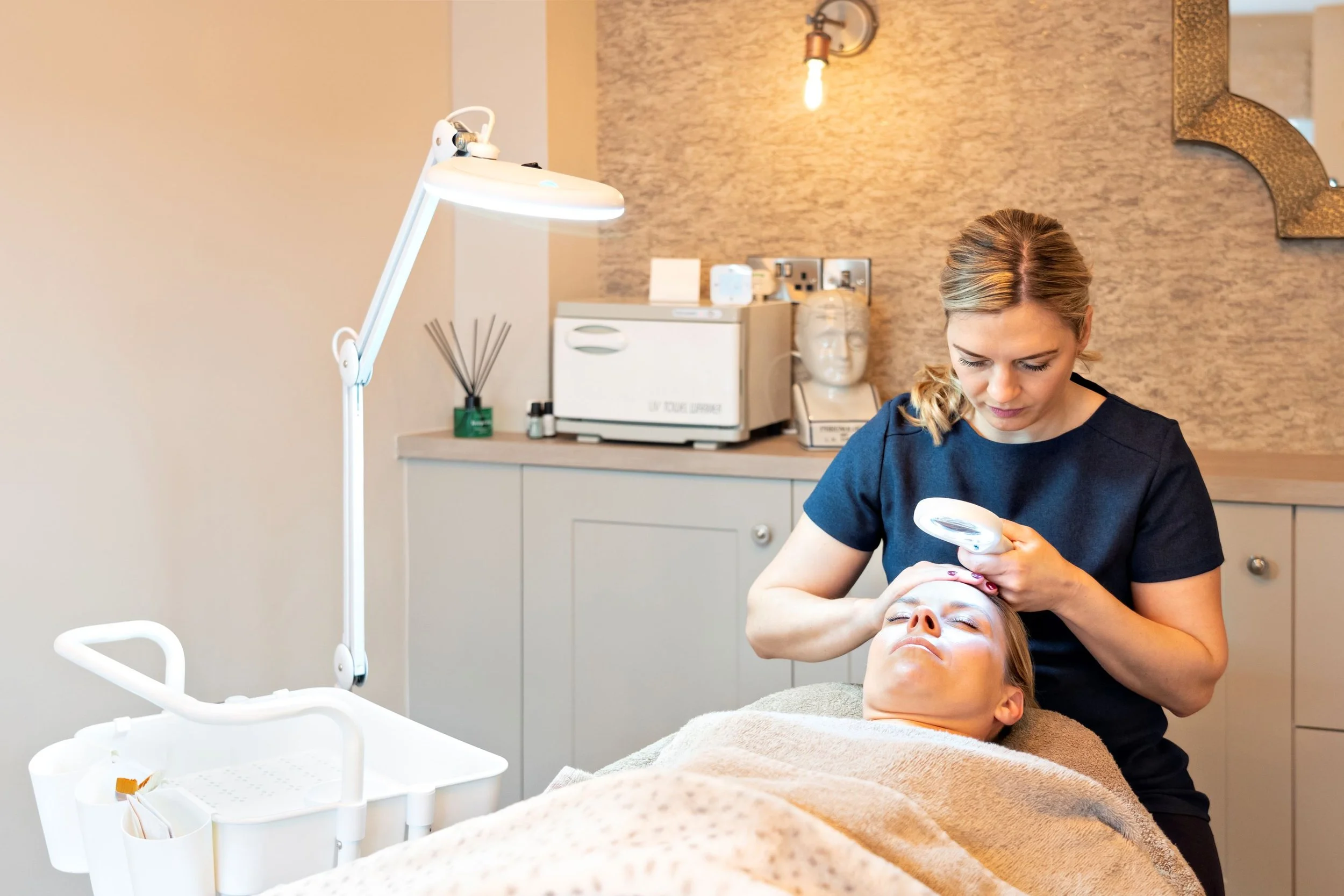 A woman receives a facial treatment from a skincare professional in a spa or clinic setting, with skincare equipment and decor visible in the background.