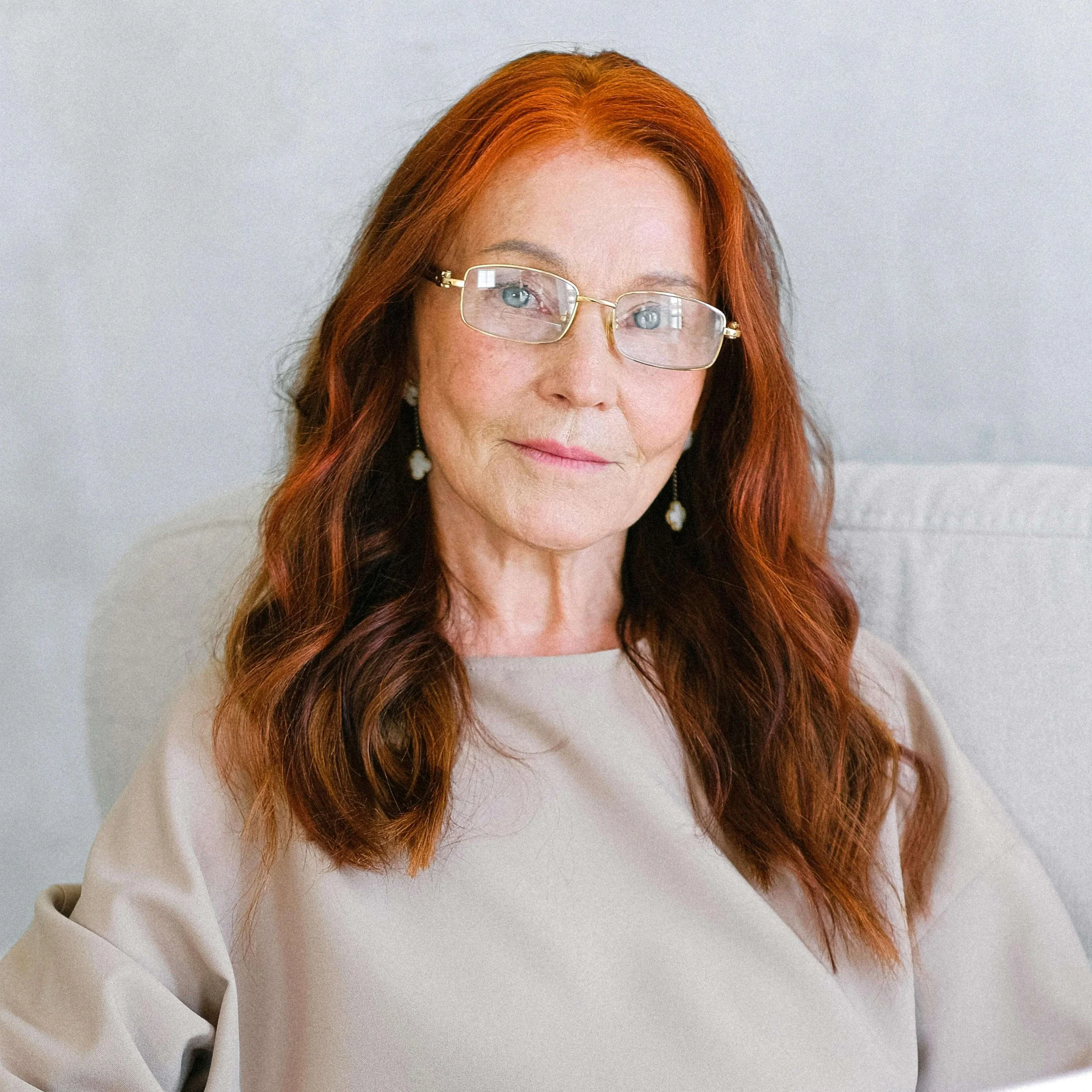 A middle-aged woman with long red hair, wearing glasses and earrings, sitting on a light-colored sofa or chair against a plain, light background.