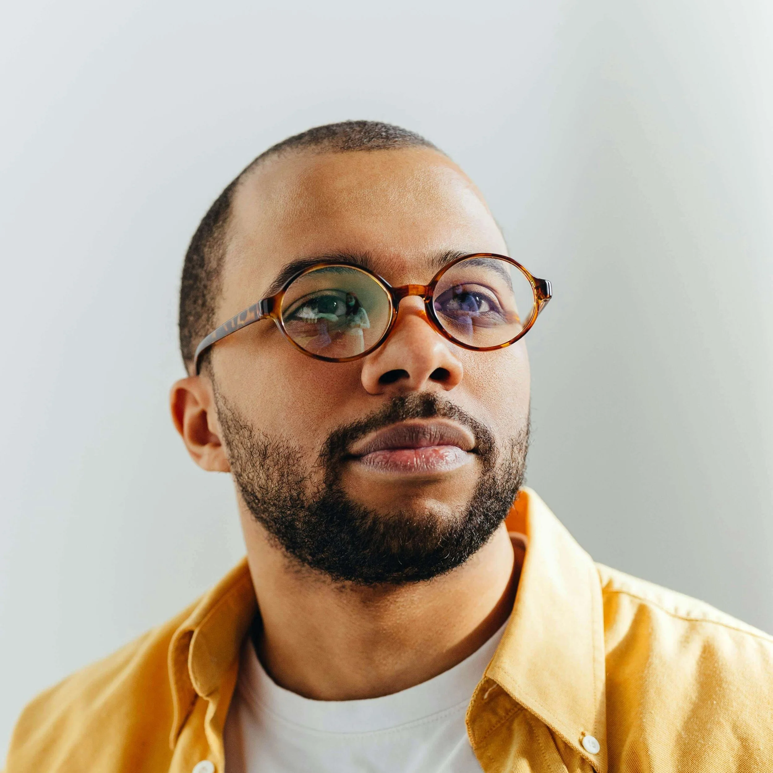 Close-up of a man with glasses, beard, and mustache, wearing a yellow shirt, against a plain background.