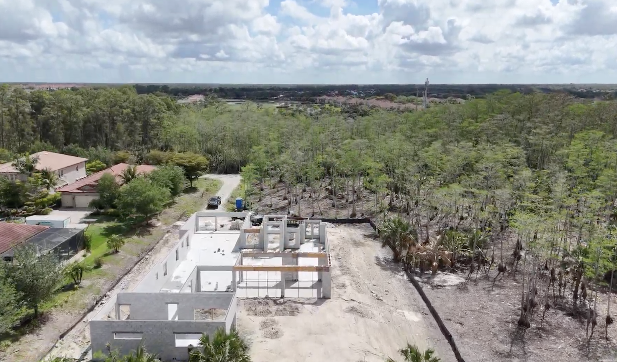 Aerial view of a residential construction site with a partially built house, surrounded by trees and neighboring homes, under a cloudy sky.