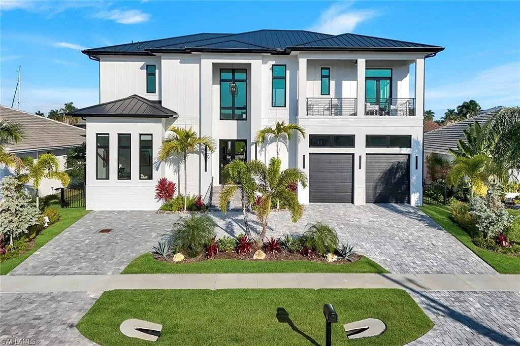 Modern two-story house with white exterior, black roof, and two garage doors, surrounded by tropical landscaping and paved driveway.