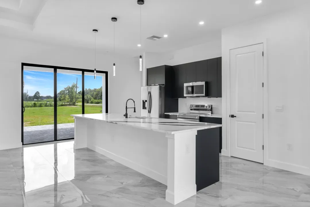 Modern kitchen with black cabinets, stainless steel refrigerator, microwave, oven, white countertops, island with sink, and sliding glass door leading outside to a grassy yard.