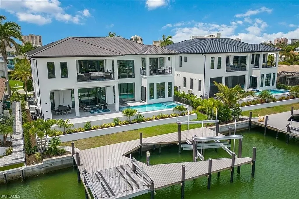 Two modern luxury houses with water views, balconies, and pools, situated along a canal with a wooden dock in a sunny suburban area.