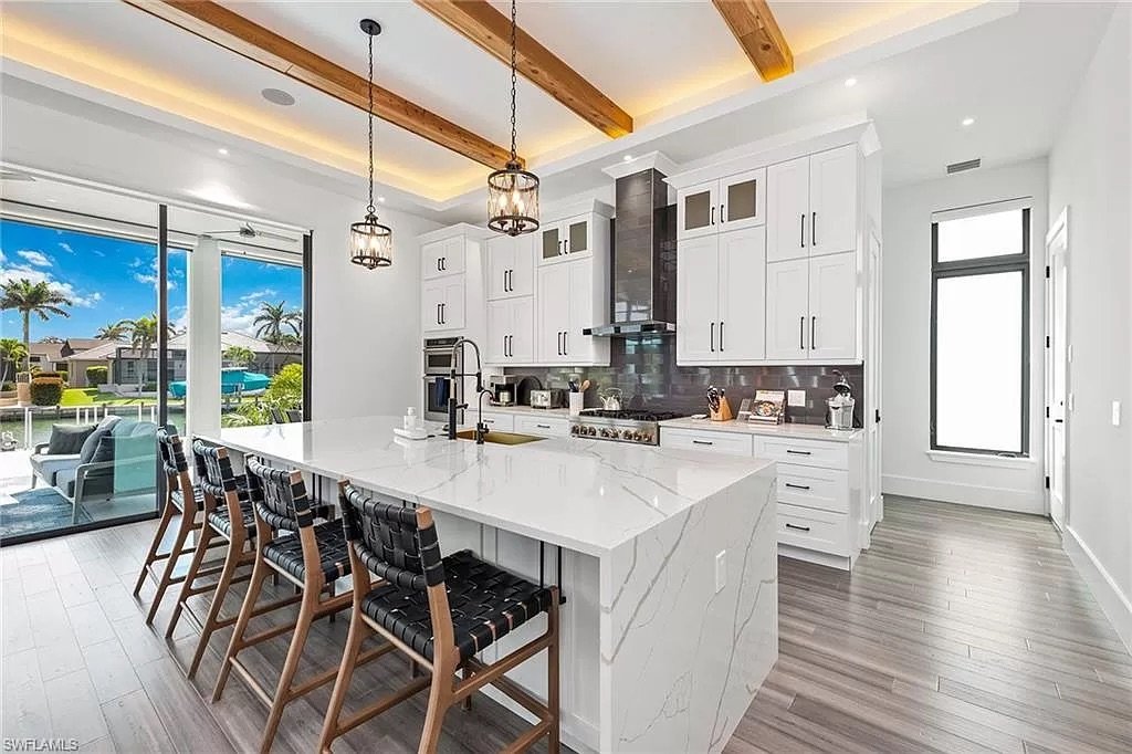 Modern kitchen with white cabinetry, marble island countertop, black backsplash, stainless steel appliances, wooden bar stools, and sliding glass door leading to patio with outdoor seating and pool overlook.