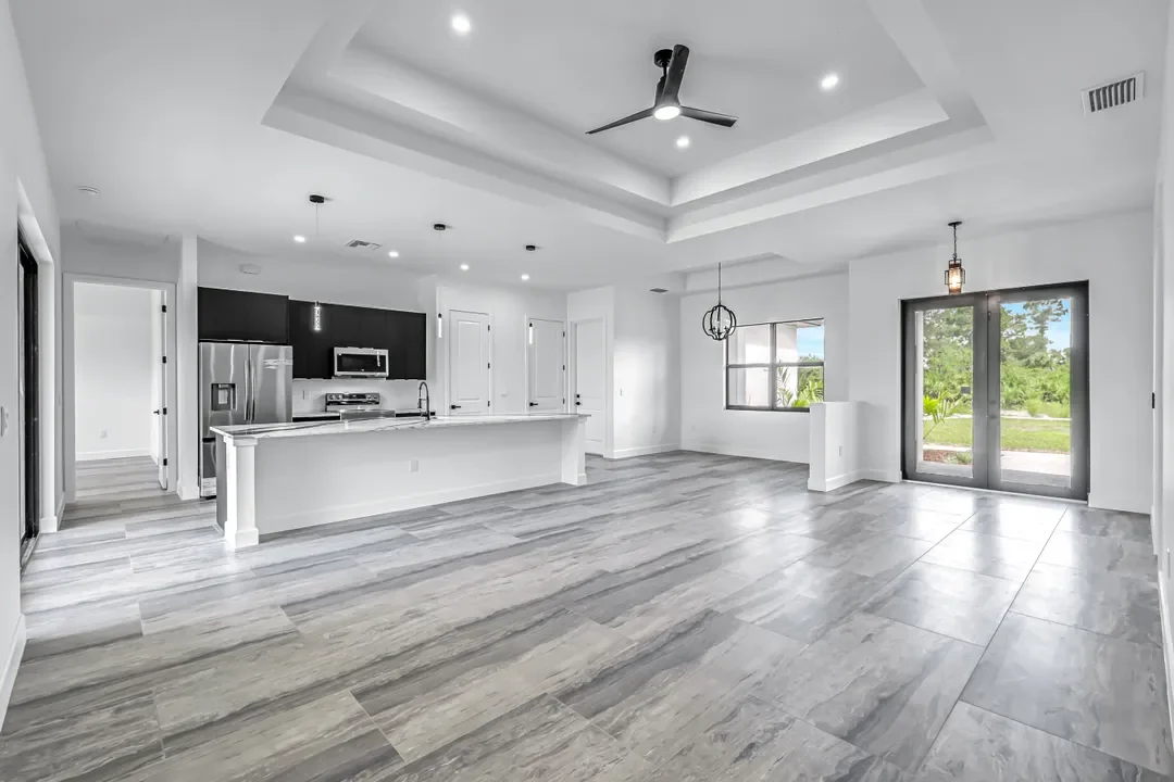 Empty modern open-concept living space with white walls, gray wood floors, a ceiling fan, kitchen with black cabinets and stainless steel appliances, large windows, and glass double doors leading outside.