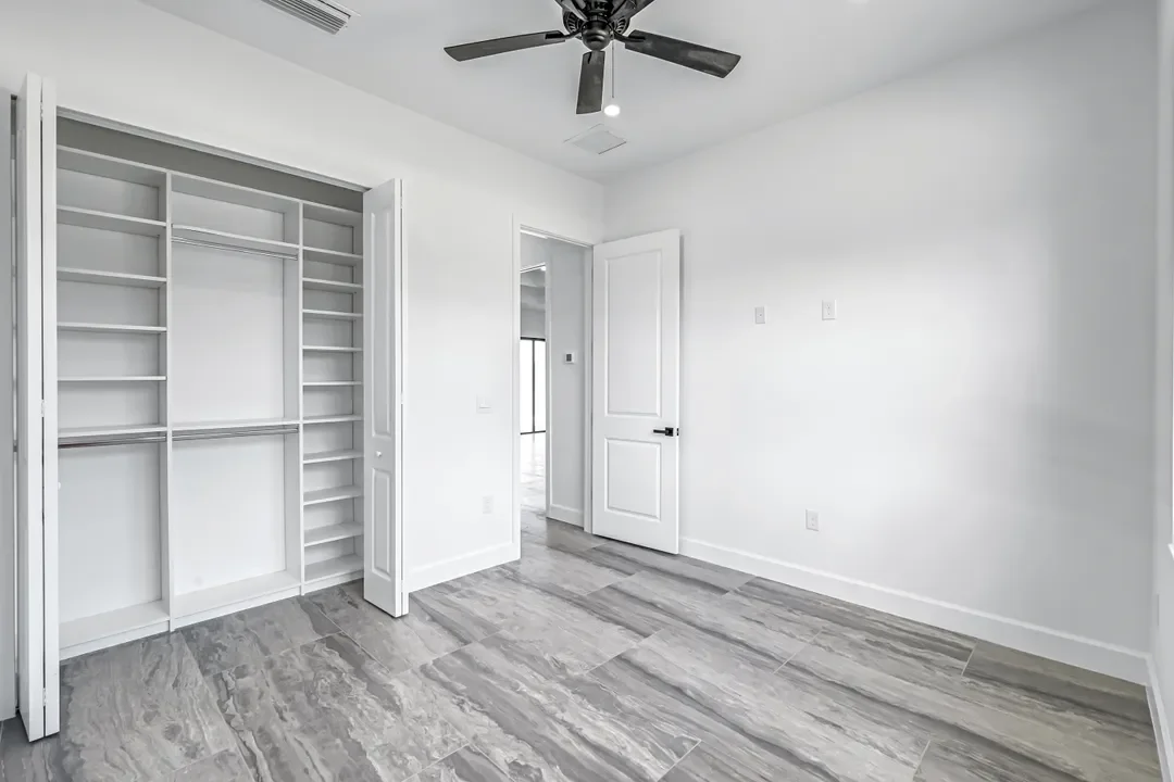 A minimalistic bedroom with white walls, an open closet with shelving, gray tile flooring, a ceiling fan, and an open door leading to another room.