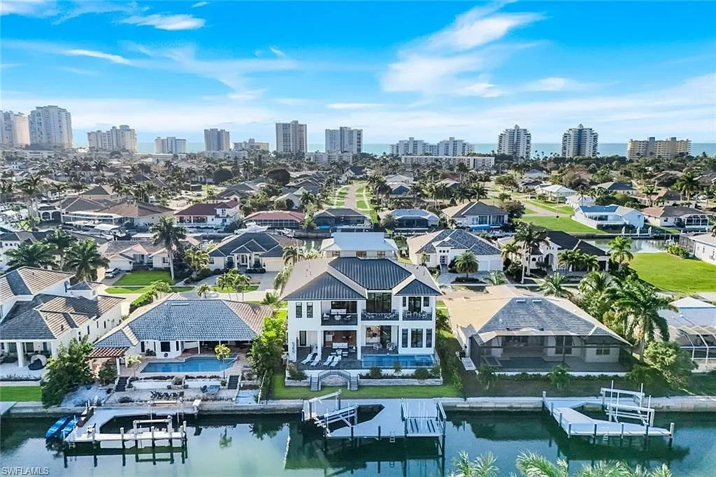 Aerial view of a waterfront residential neighborhood with houses, palm trees, and docks along a canal, with high-rise buildings in the background under a partly cloudy sky.