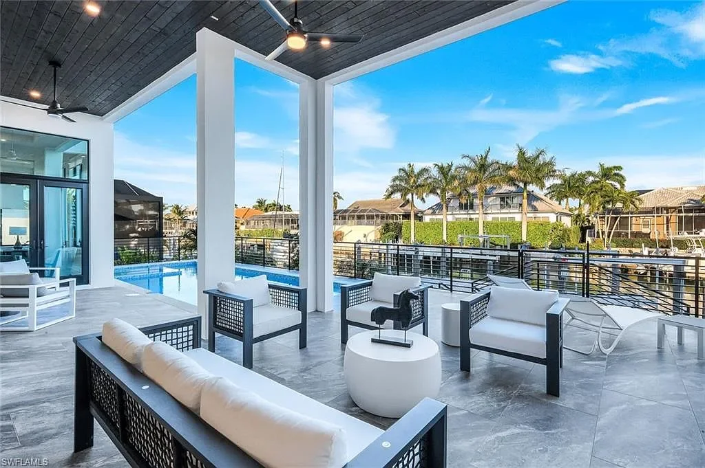 Patio area with white and black outdoor furniture, overlooking a swimming pool and backyard with palm trees and houses under a blue sky.