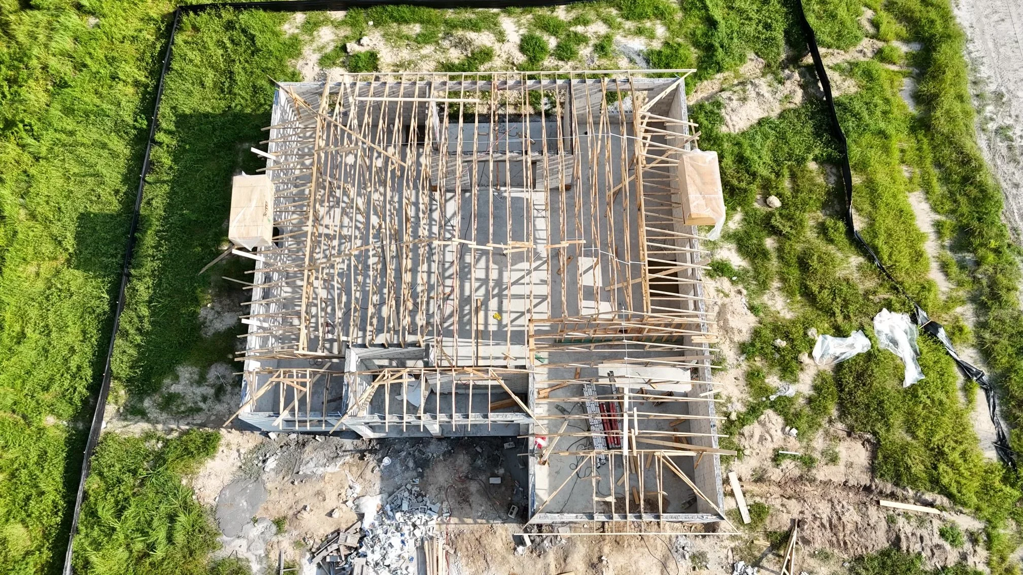 Aerial view of a house under construction with visible wooden framing, surrounded by green vegetation and construction materials.