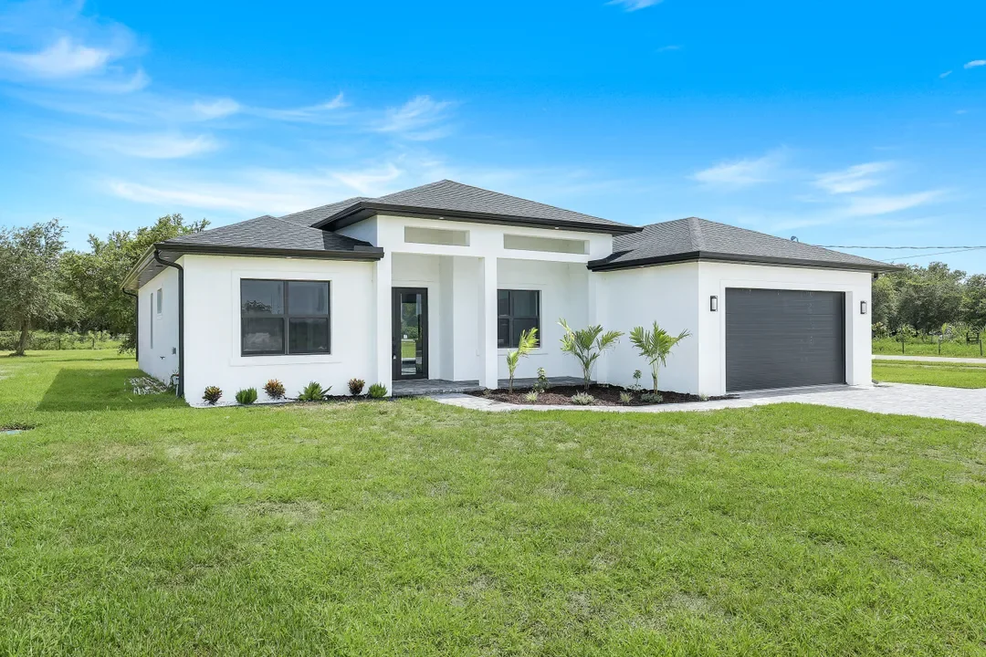 Front view of a modern white house with a gray roof, black garage door, and small front garden with palm trees, on a sunny day with blue sky.