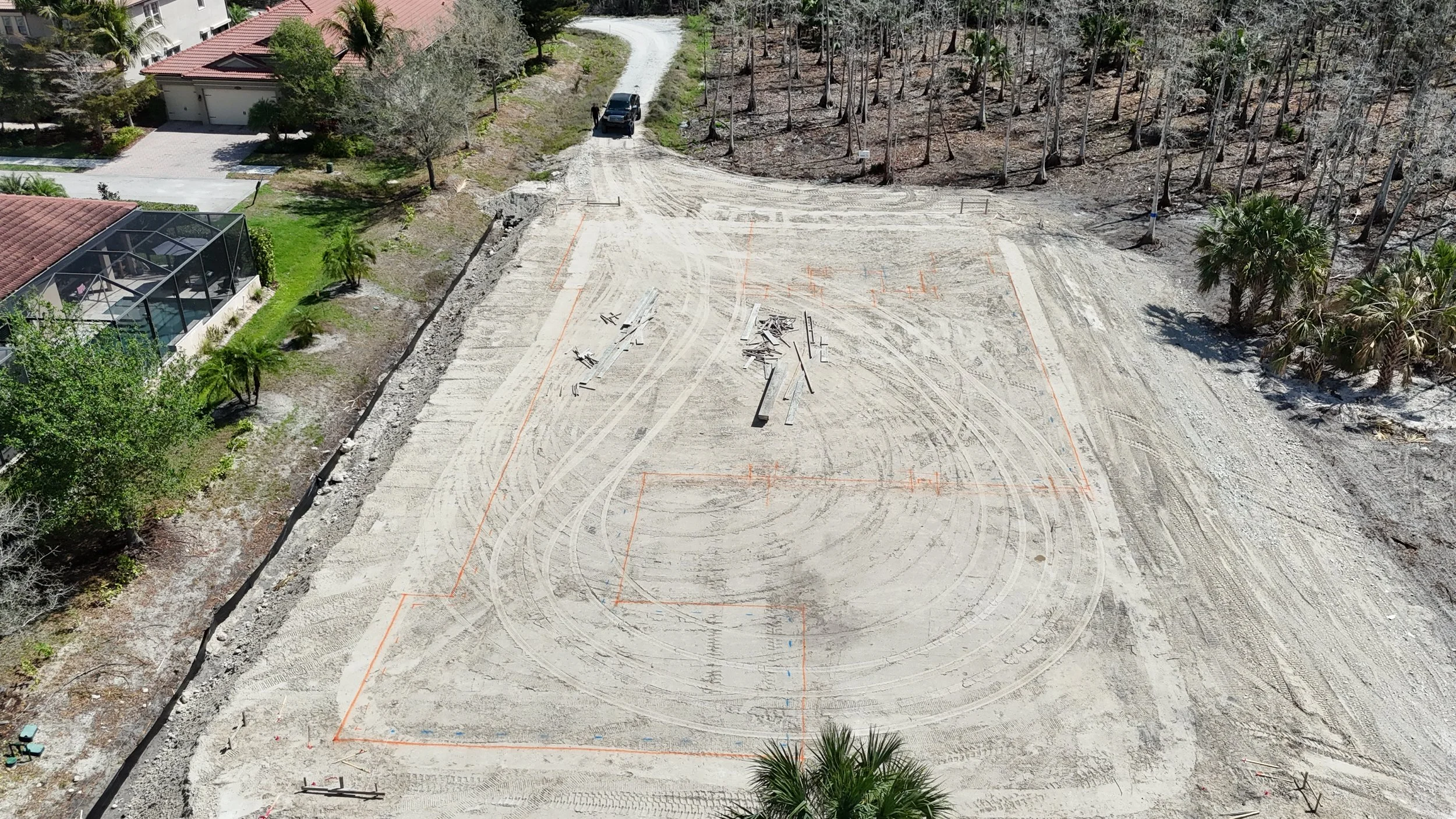 A construction site with cleared land and orange boundary lines, surrounded by trees and residential houses, with construction materials and tire tracks visible on the dirt.