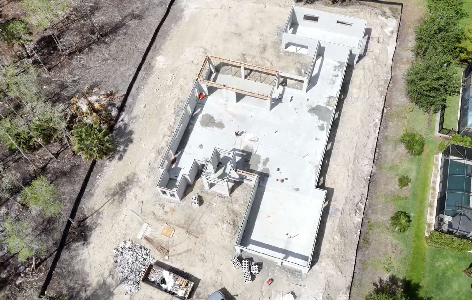 Aerial view of a house under construction, showing its concrete foundation and framing in progress, surrounded by trees and neighboring properties.