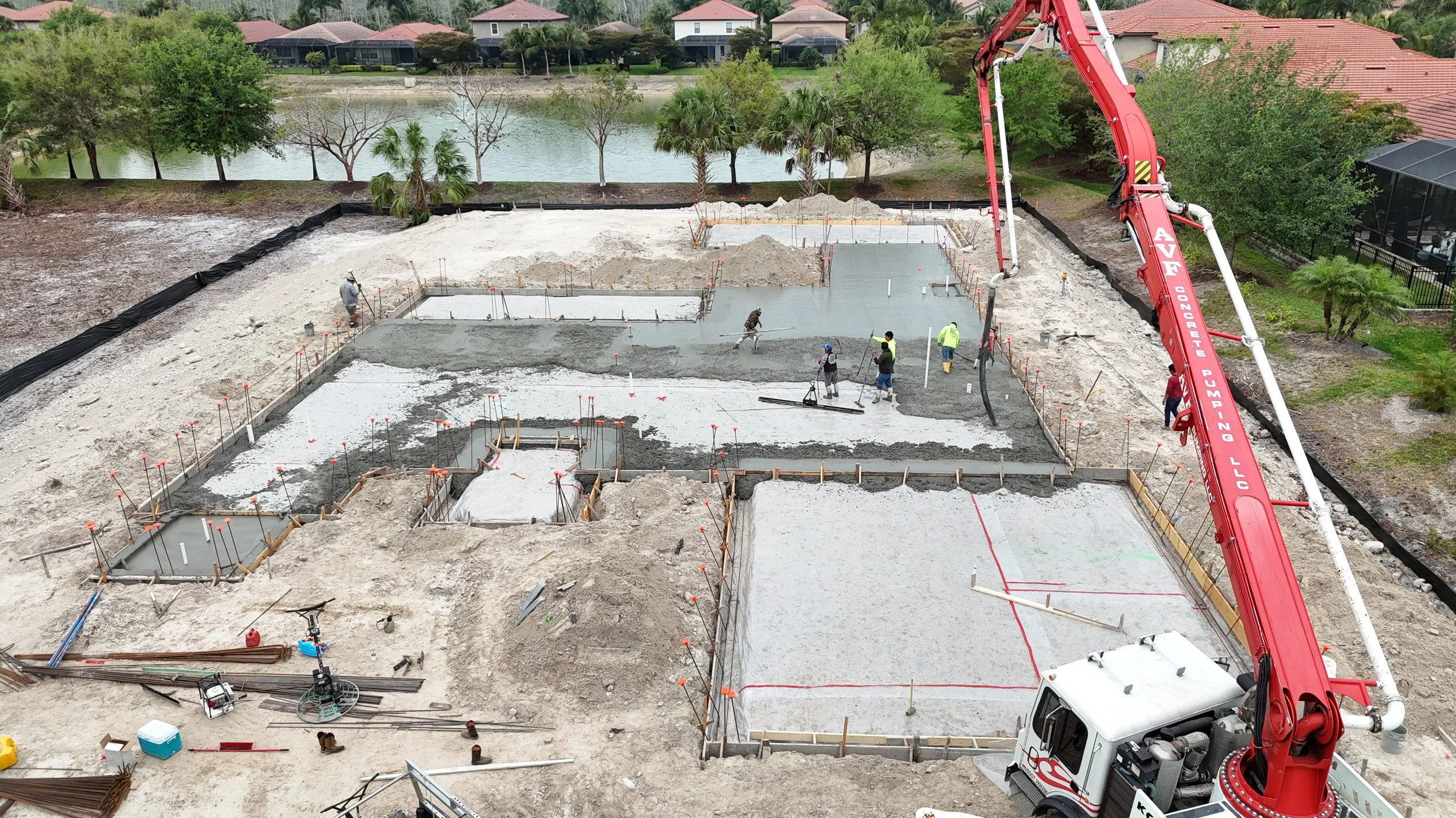 Construction workers pouring concrete for a building foundation near a lake, with a red concrete pump truck on site.
