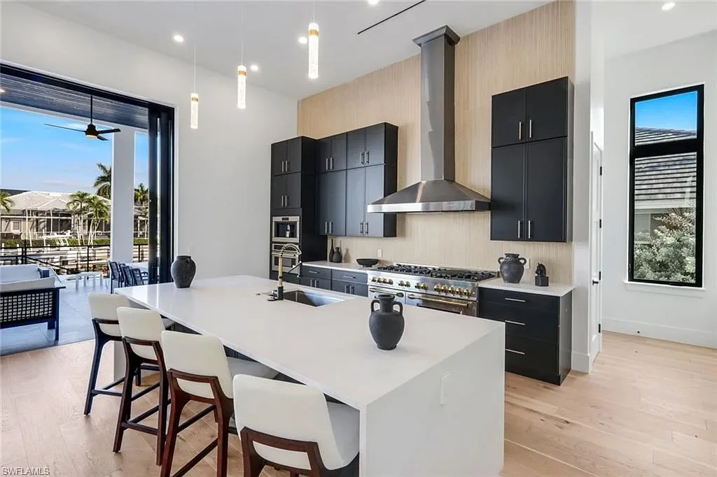 Modern kitchen with white island, black cabinets, stainless steel range with vent hood, and large window displaying outdoor patio and neighborhood.