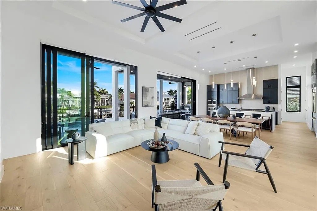 Bright open-concept living room and kitchen with large windows, white sectional sofa, modern black ceiling fan, light wood flooring, and minimal decor.