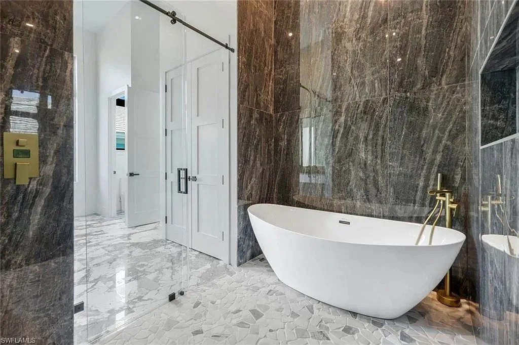 Modern bathroom with a white bathtub, gold fixtures, marble wall, and marble floor. Glass shower door and white doors in the background.