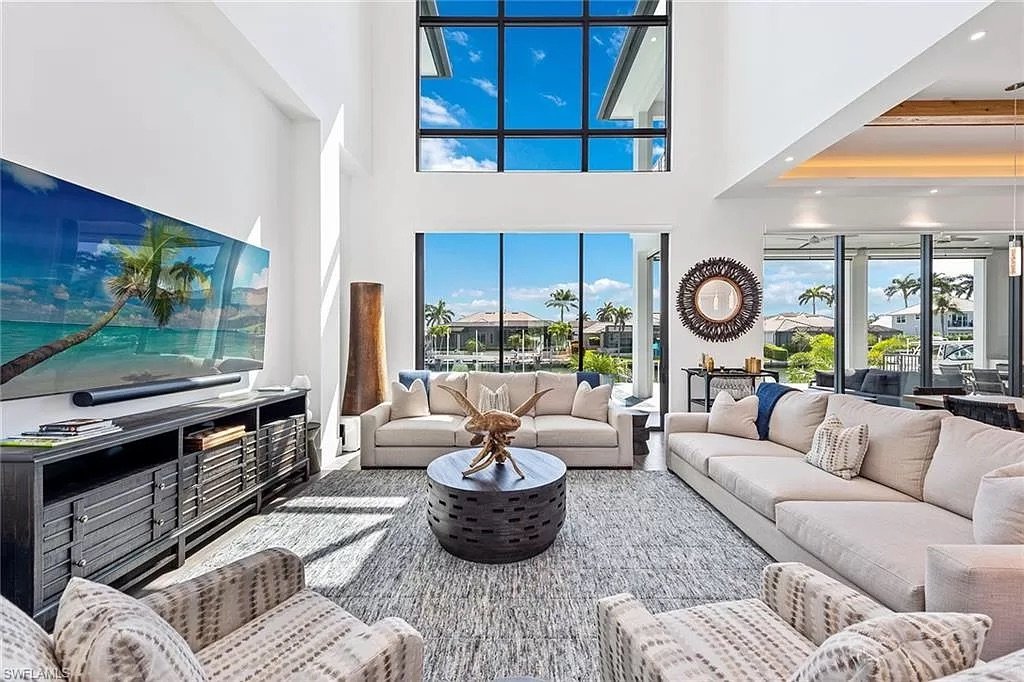 Bright modern living room with high vaulted ceilings, large windows showing a view of the ocean and palm trees, beige sofas, and a large flatscreen TV.