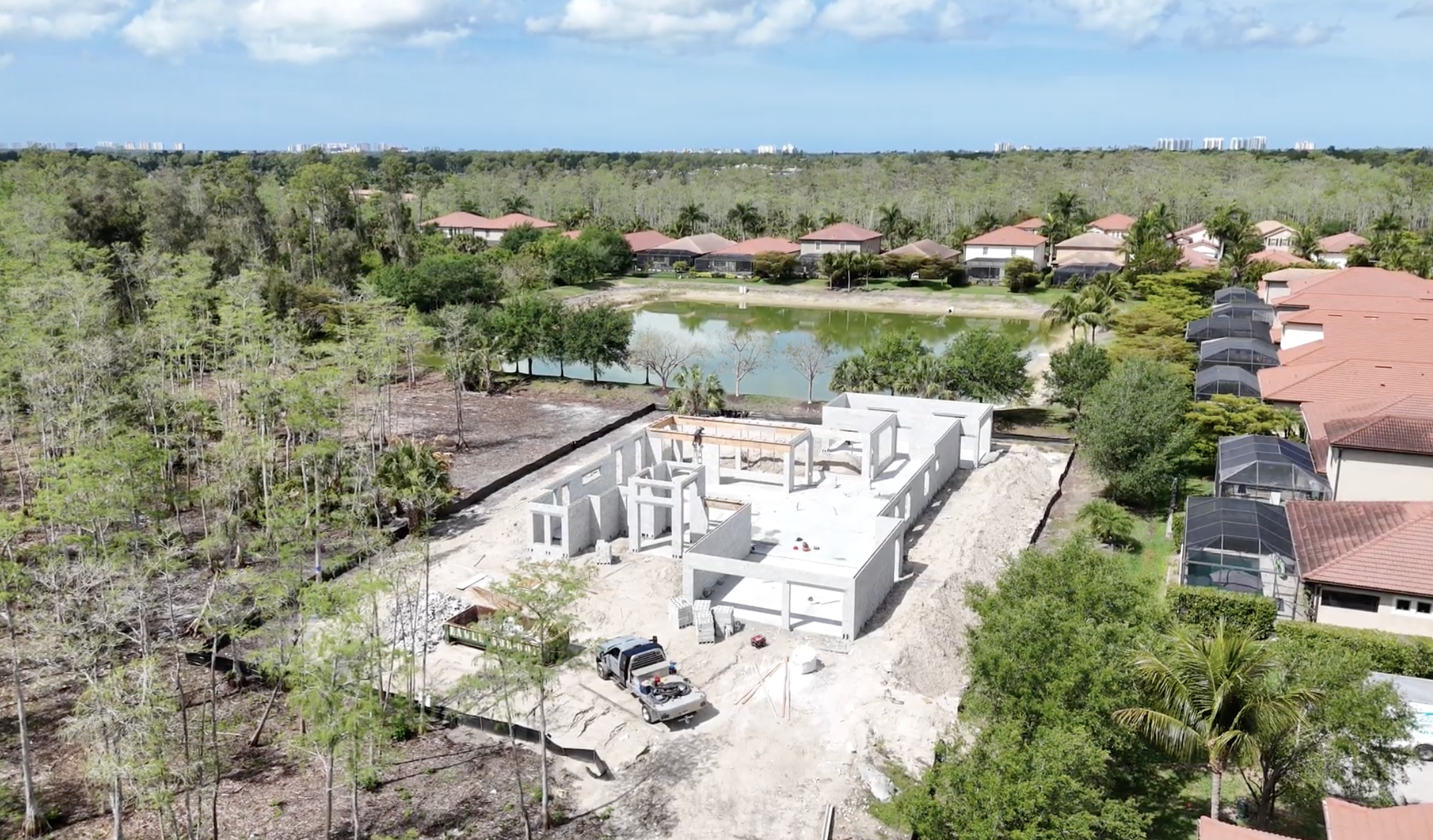 Aerial view of a house under construction in a neighborhood with a pond and trees in the background.
