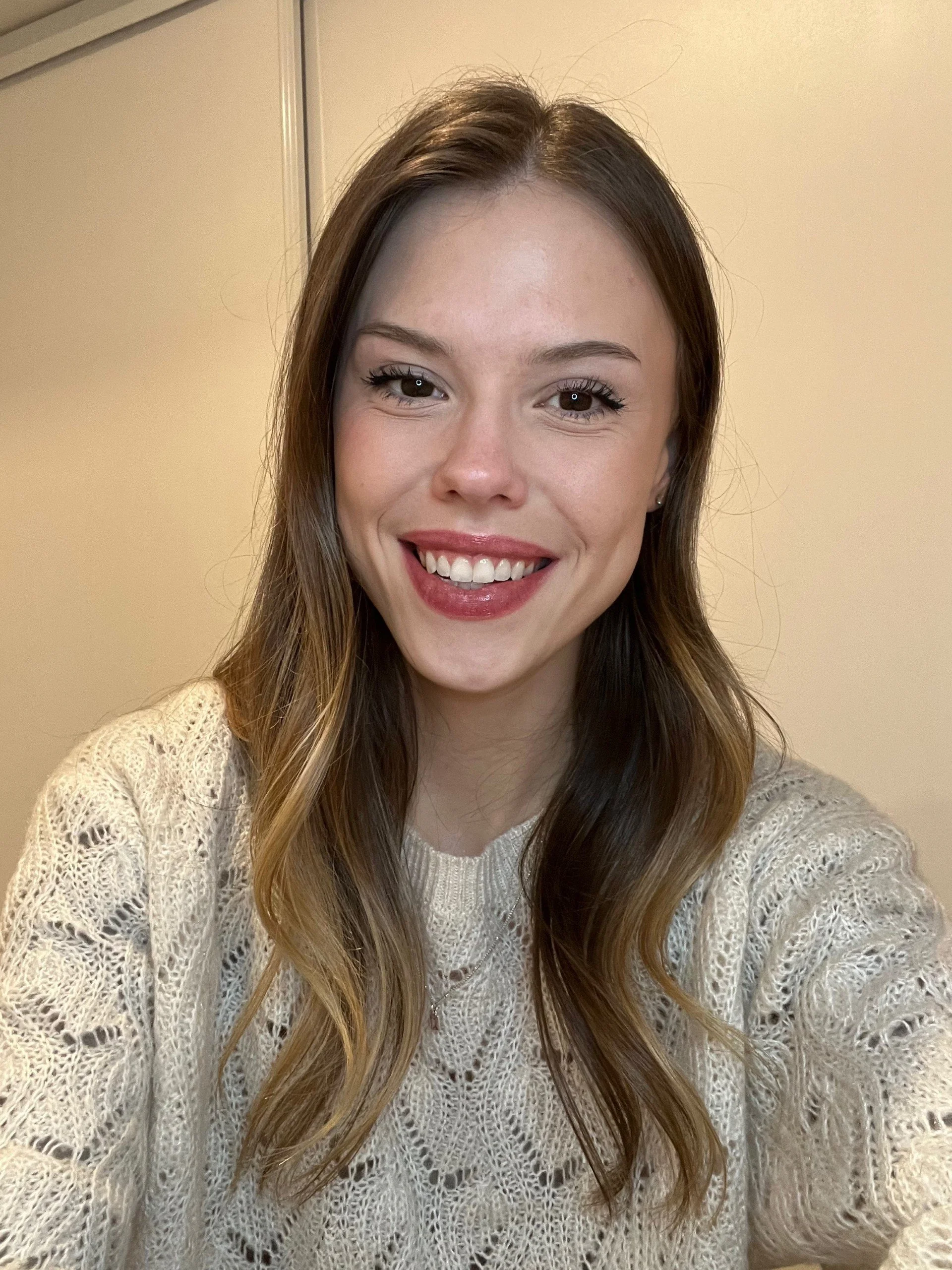 A young woman with shoulder-length wavy brown hair taking a selfie indoors. She is smiling, showing braces, and wearing a cream-colored knit sweater.
