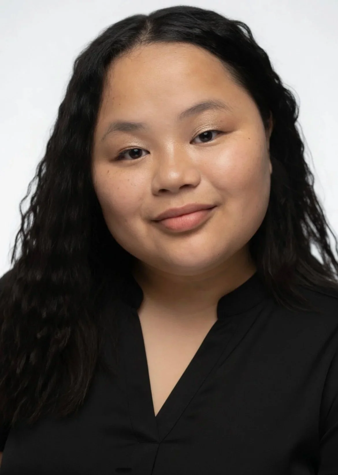 Close-up of a woman with long, wavy black hair, fair skin, and wearing a black top, smiling softly against a plain white background.