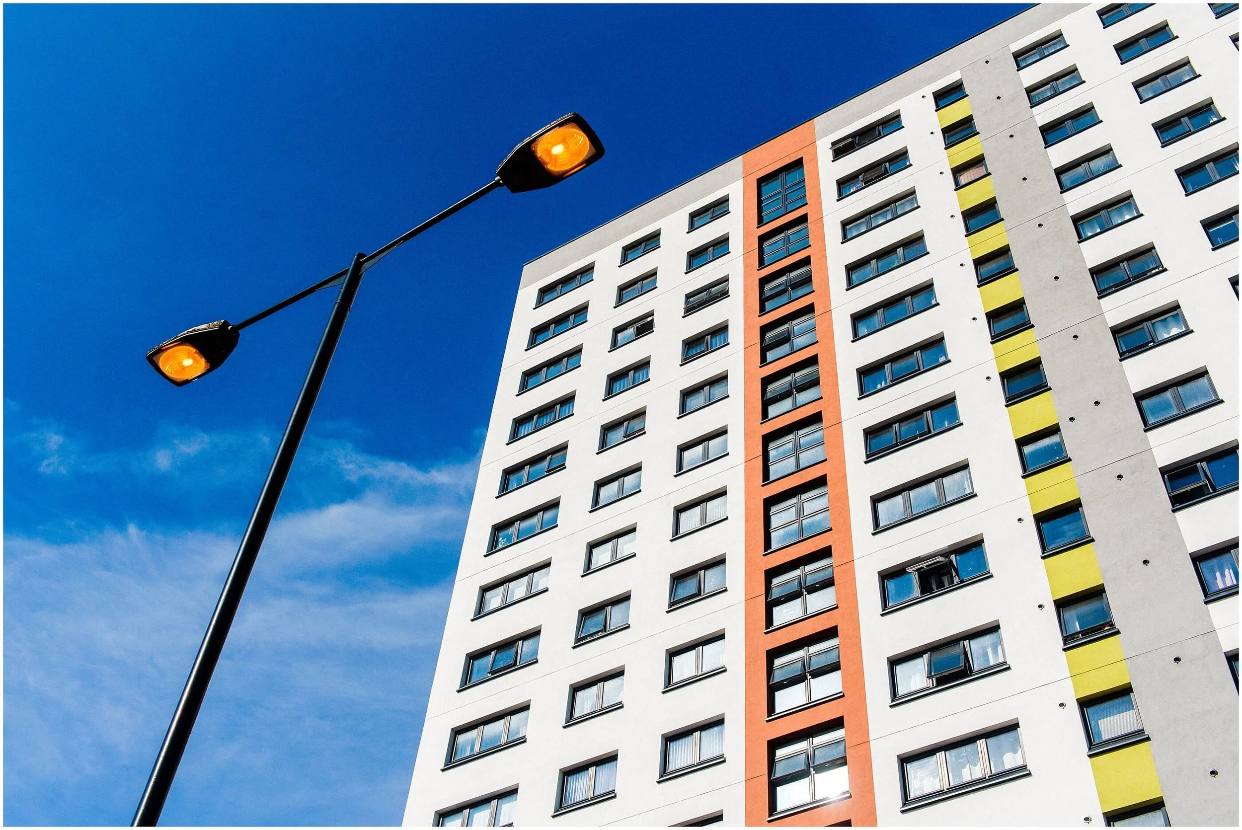 A tall streetlamp with two orange lights against a modern multi-story building with colorful accents and multiple windows, under a clear blue sky.